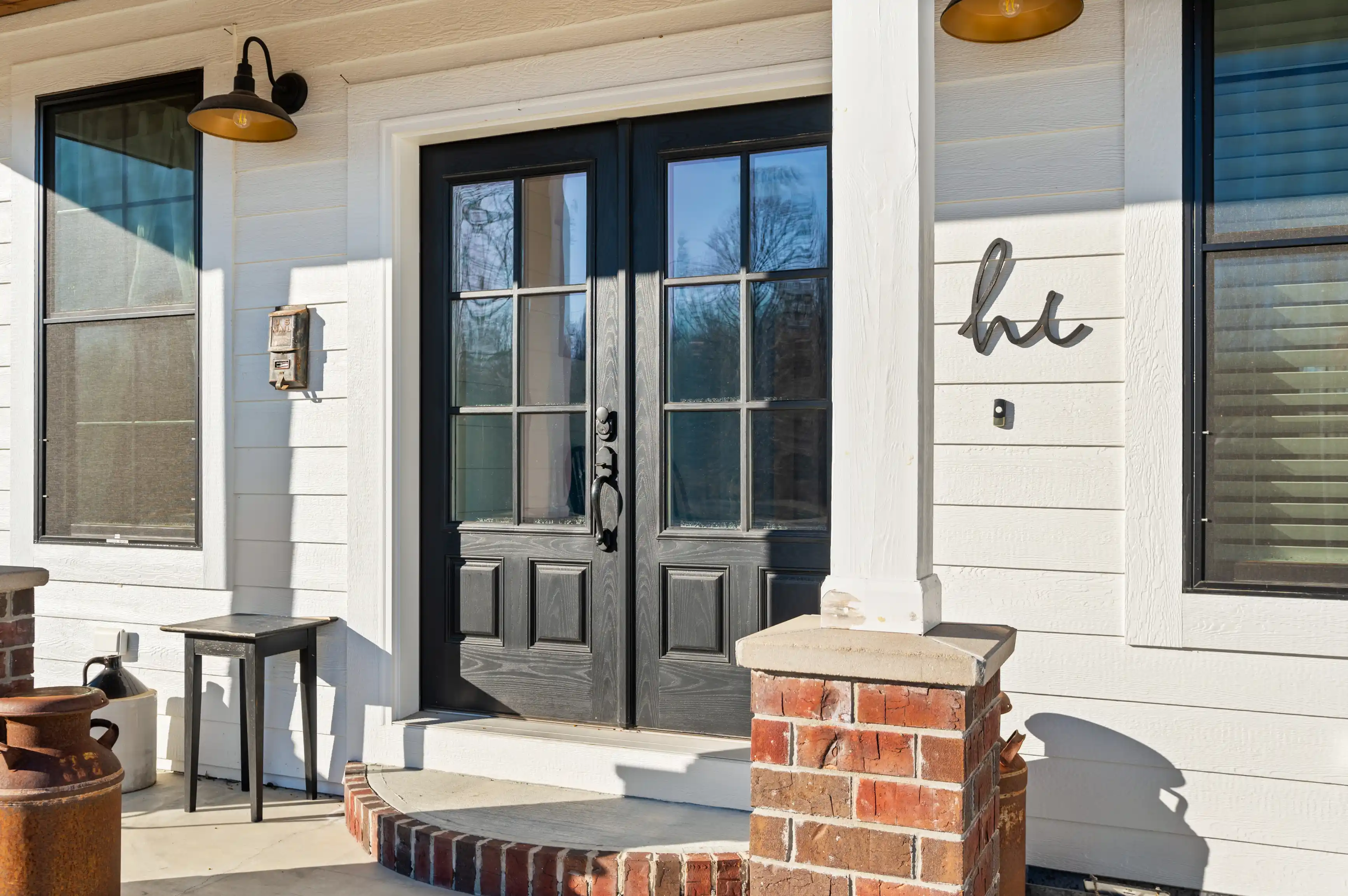 Front porch of a house with black double doors, side windows, and "hi" sign, with a rustic lamp above and a white wall.