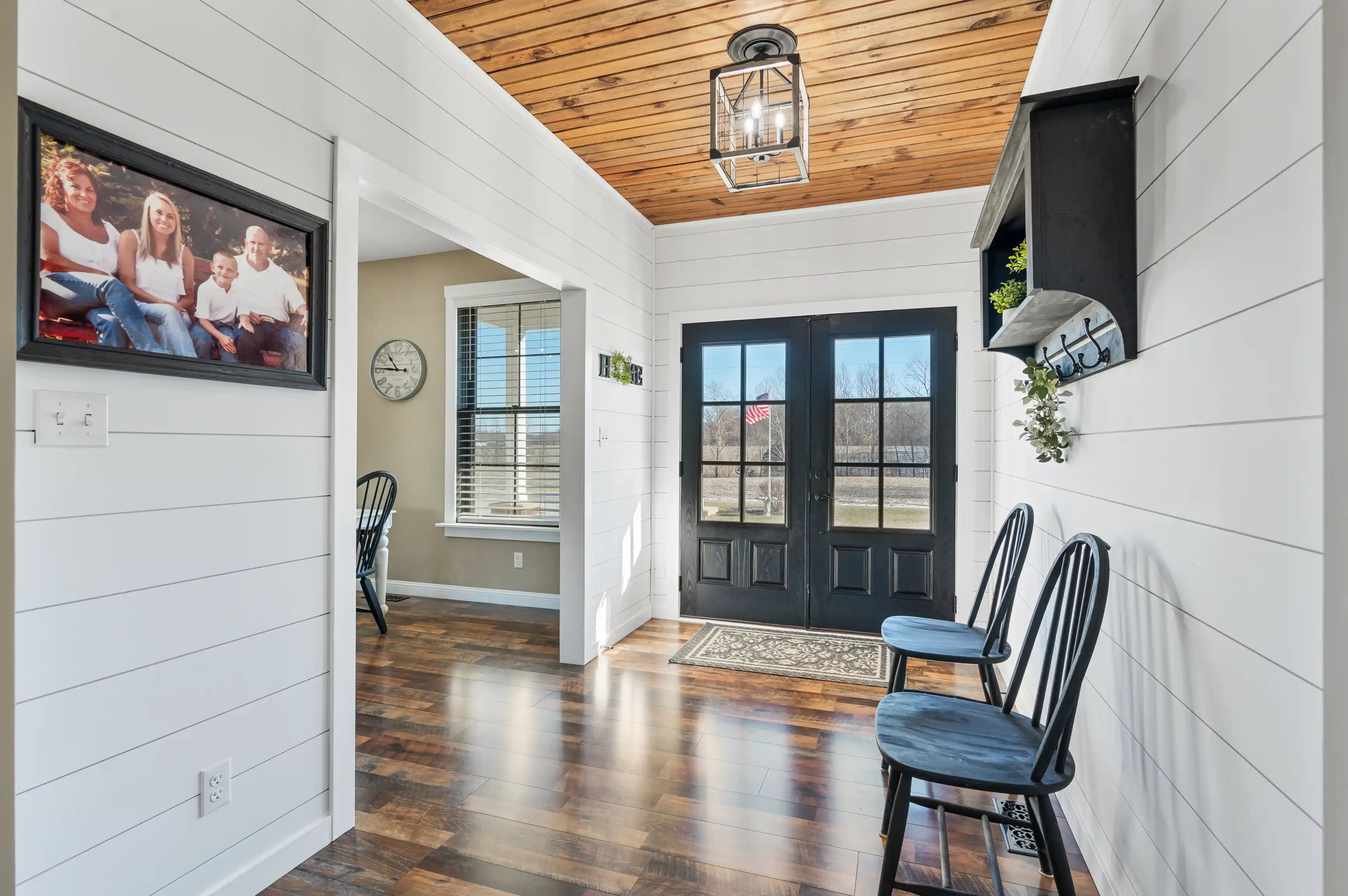 Bright interior entryway with wooden flooring, a white shiplap wall, a wooden ceiling, black double doors, and decorative elements including wall-mounted coat hooks and a framed family photograph.