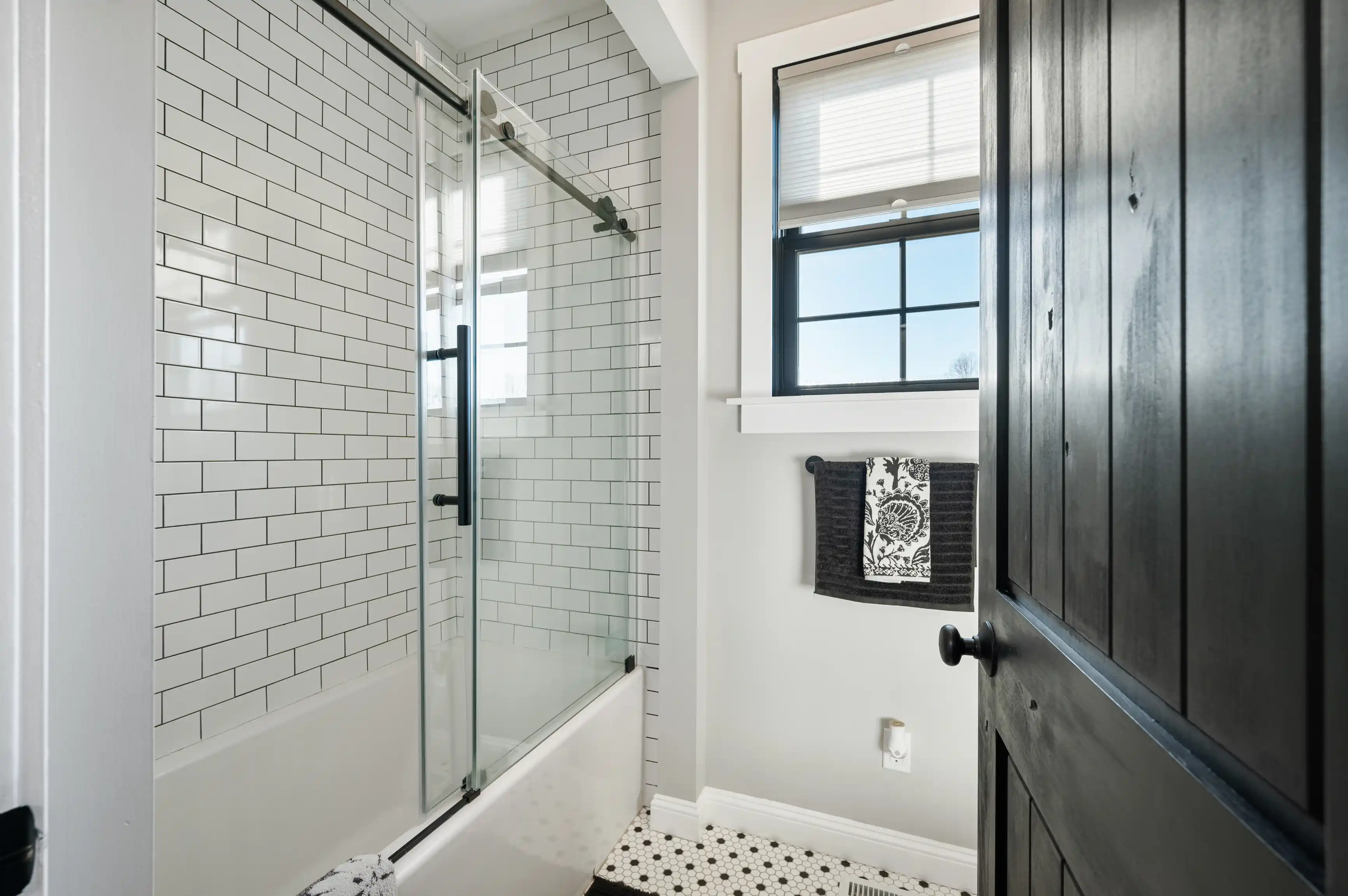 A modern bathroom with white subway tiles, glass door shower, a black framed window with blinds, patterned black and white floor tiles, and a dark wood linen closet.