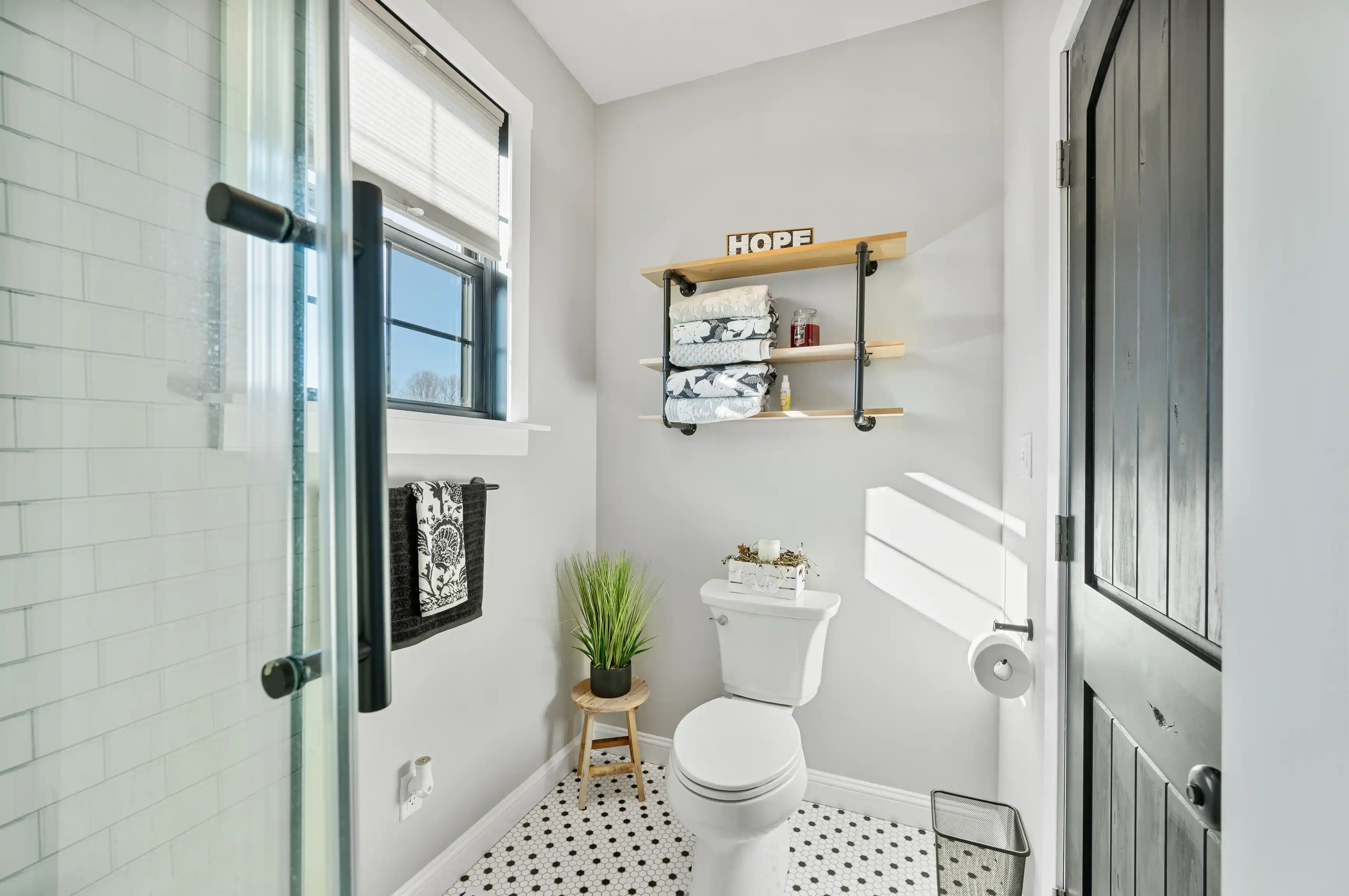Brightly lit bathroom interior with glass shower door, white subway tiles, floating wooden shelves with towels, 'HOPE' sign, toilet, and plant decor.