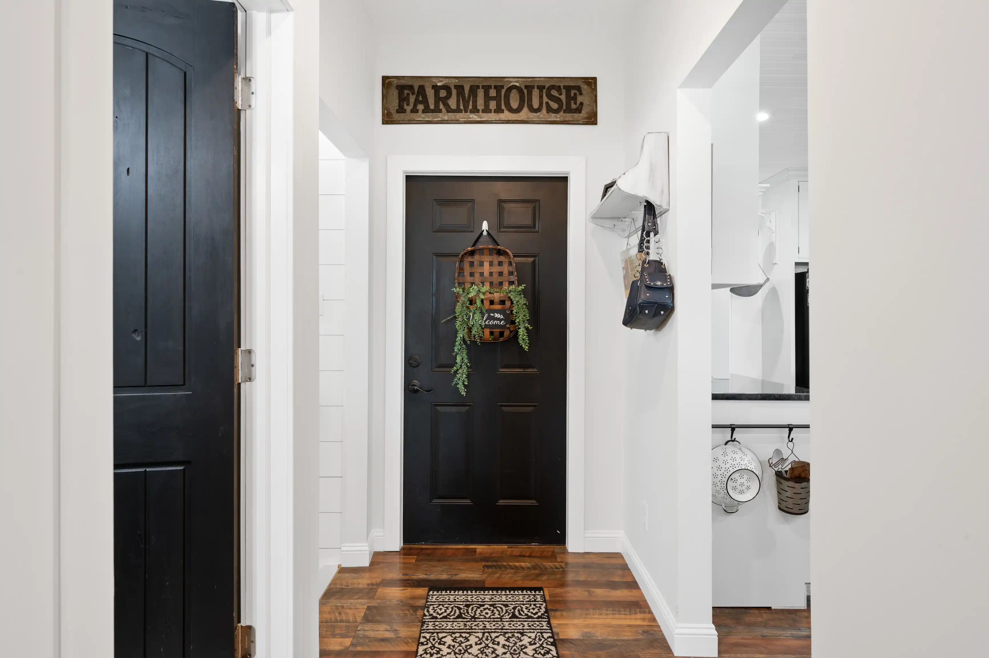 Modern farmhouse style hallway with a decorative "FARMHOUSE" sign, black front door adorned with a welcome basket and greenery, patterned rug on wooden floor, and hanging accessories on white walls.