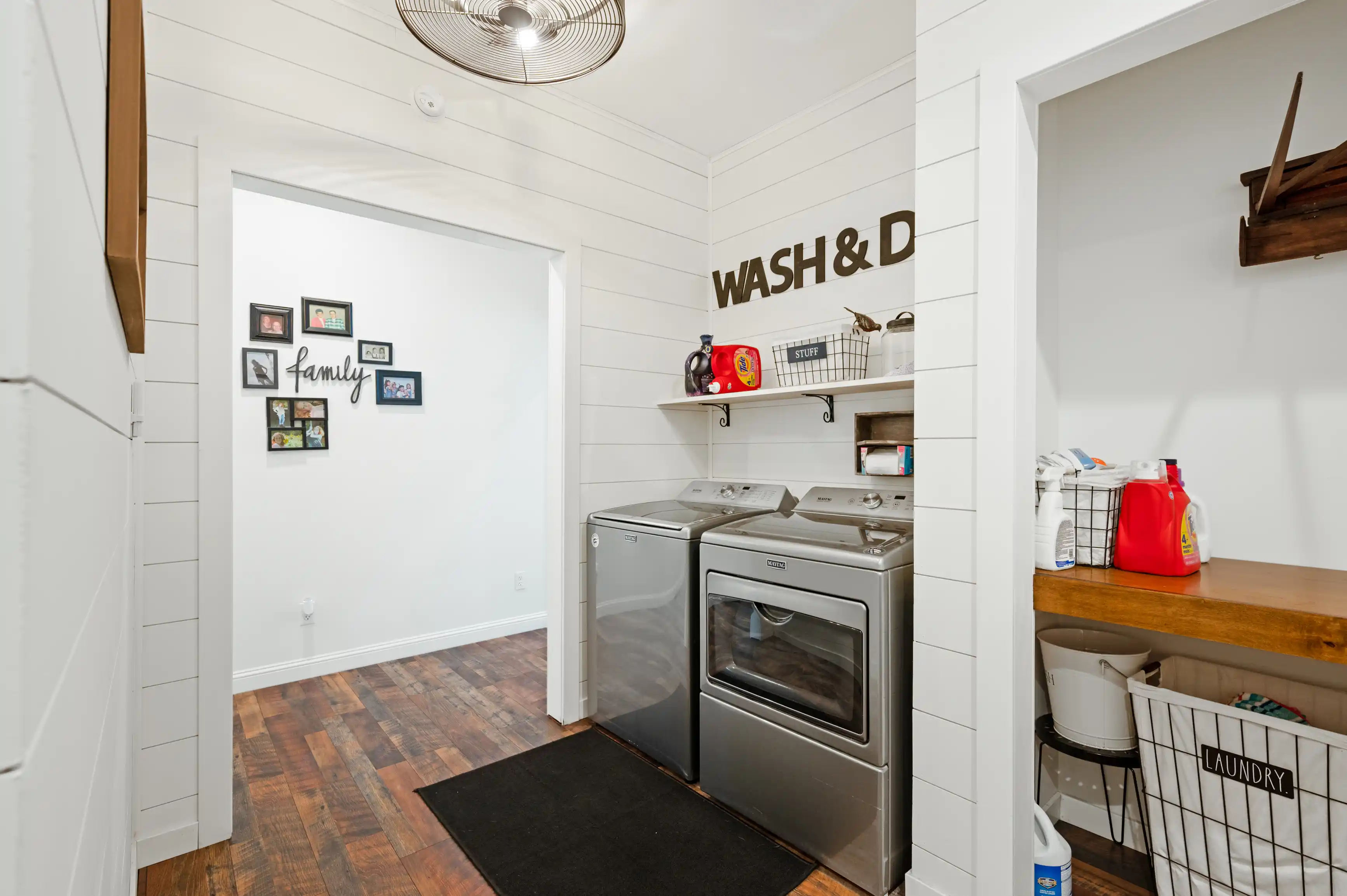 Bright and modern laundry room with white shiplap walls, stainless steel washer and dryer, wooden countertop, open shelving with supplies, and decorative 'WASH & DRY' lettering on the wall.