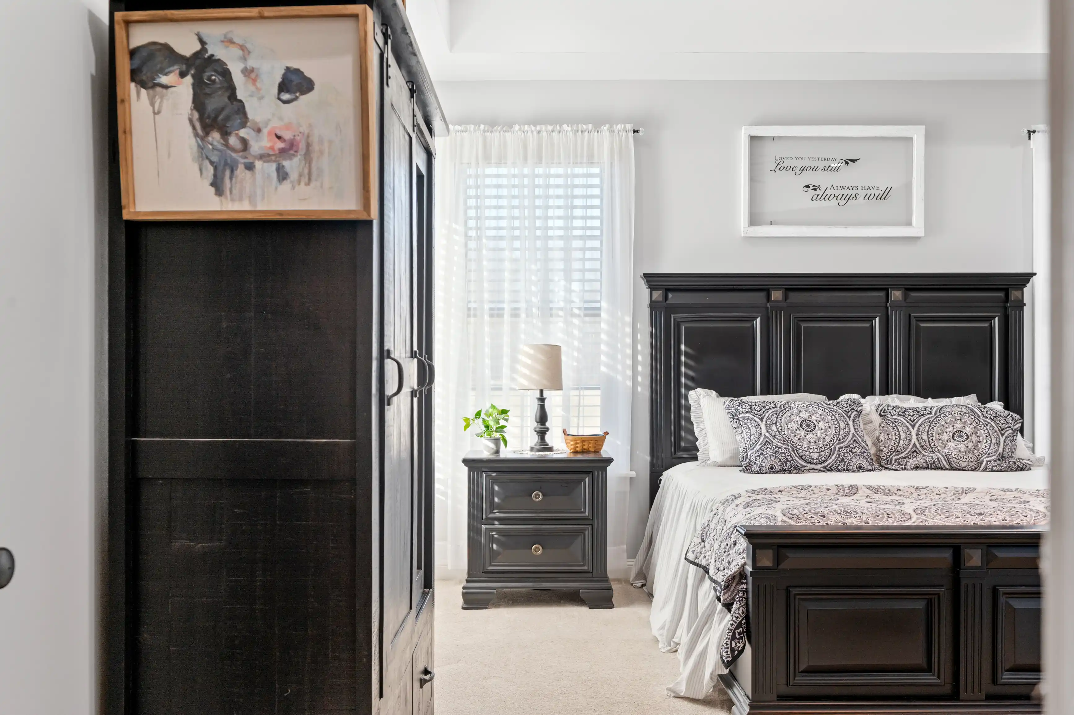 A cozy bedroom with white sheer curtains, elegant black furniture including a bed with patterned bedding, a nightstand with a lamp, and wall art above the headboard with an inspirational quote.
