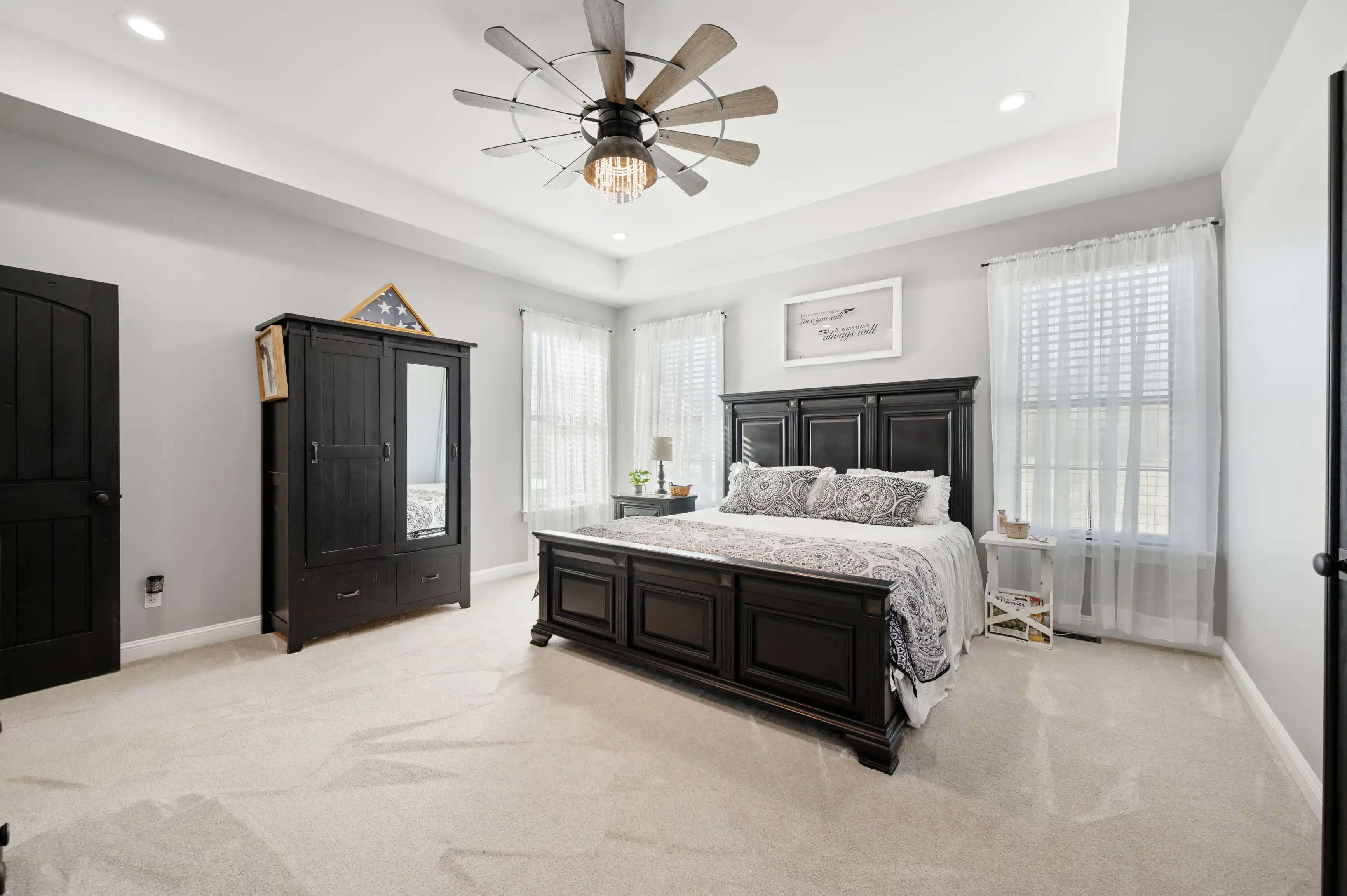 Elegant bedroom featuring a large black bed with patterned bedding, a wooden wardrobe, a modern ceiling fan, sheer curtains, and framed wall decor.