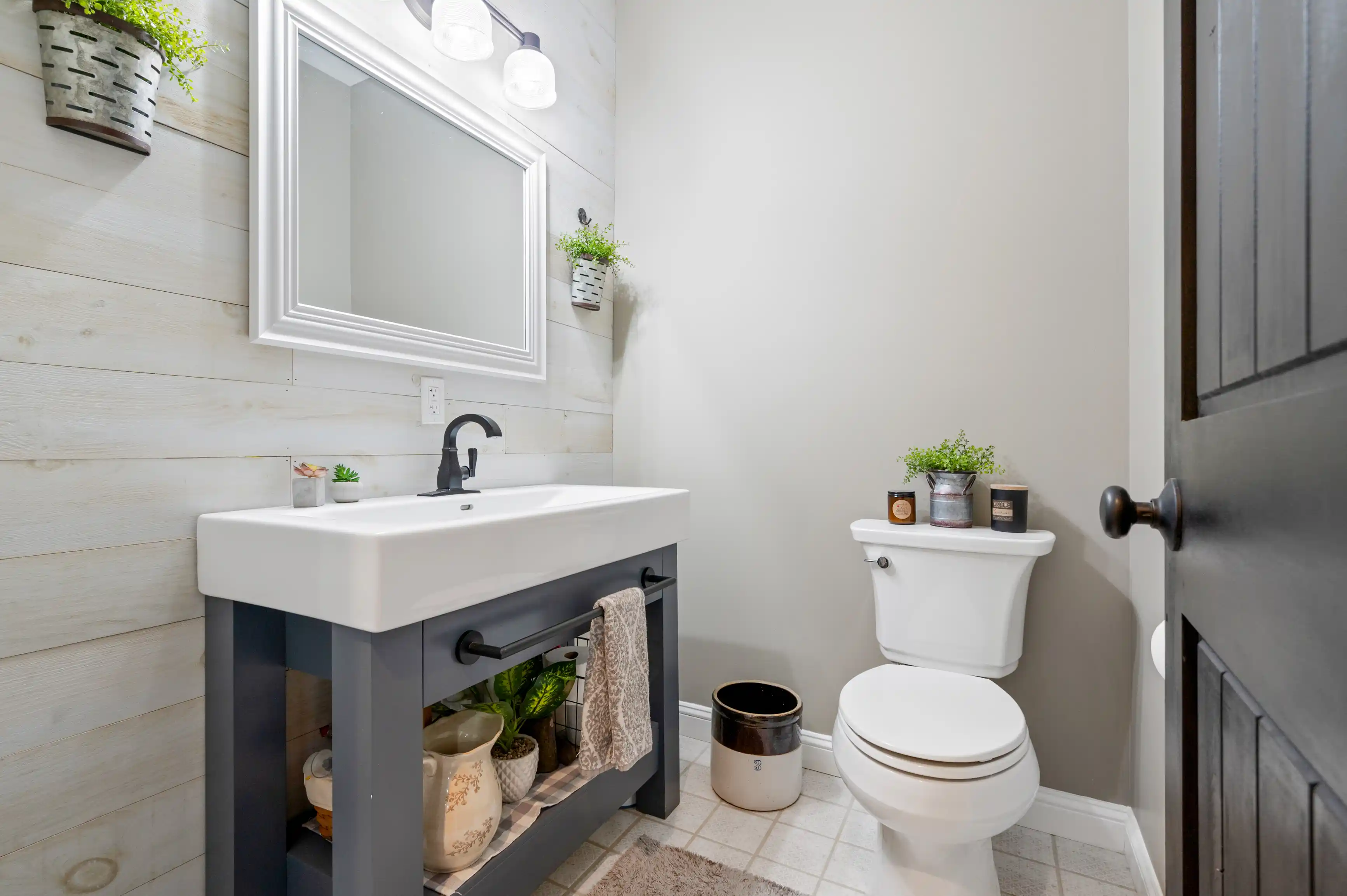 Modern bathroom interior with white shiplap walls, vanity cabinet with sink, mirror, toilet, and decorative plants.