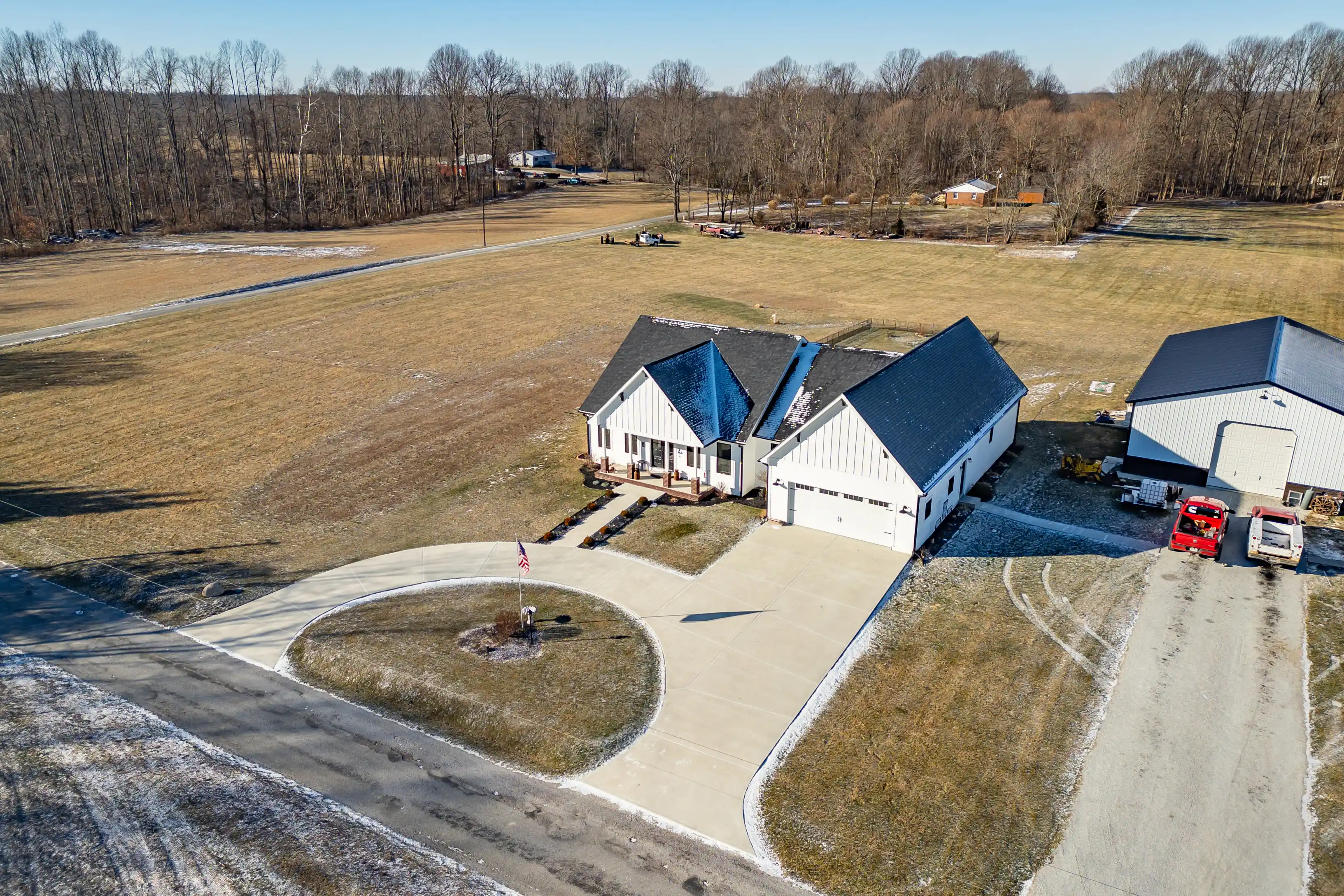Aerial view of a rural residential property with a white house and separate garage, circular driveway, and surrounding grassy fields, with trees in the background.