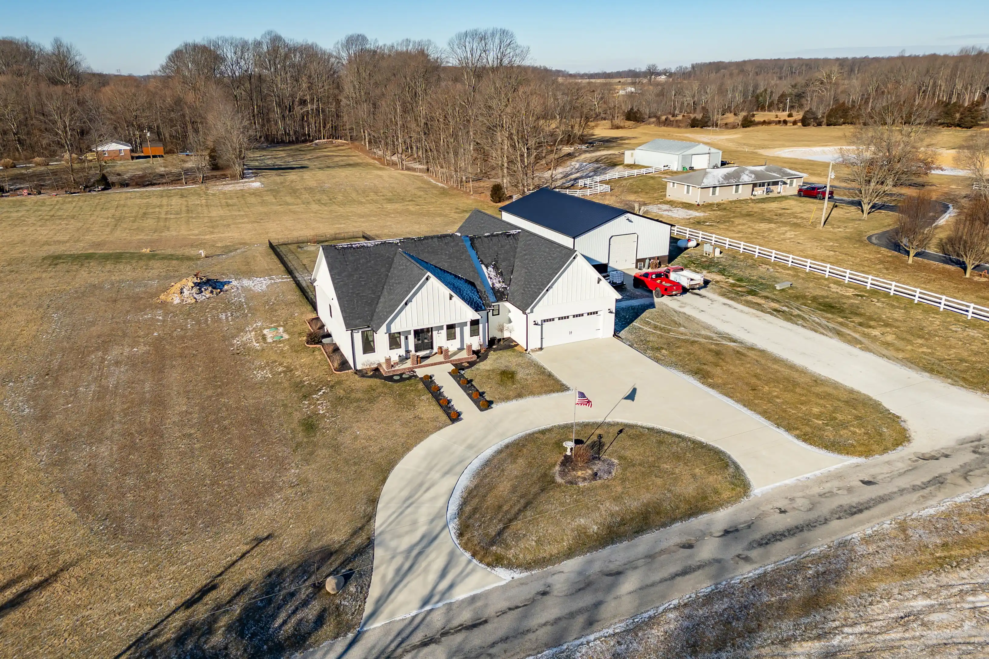 Aerial view of a residential property with a large white house, attached garage, circular driveway, American flag, and surrounding open land.