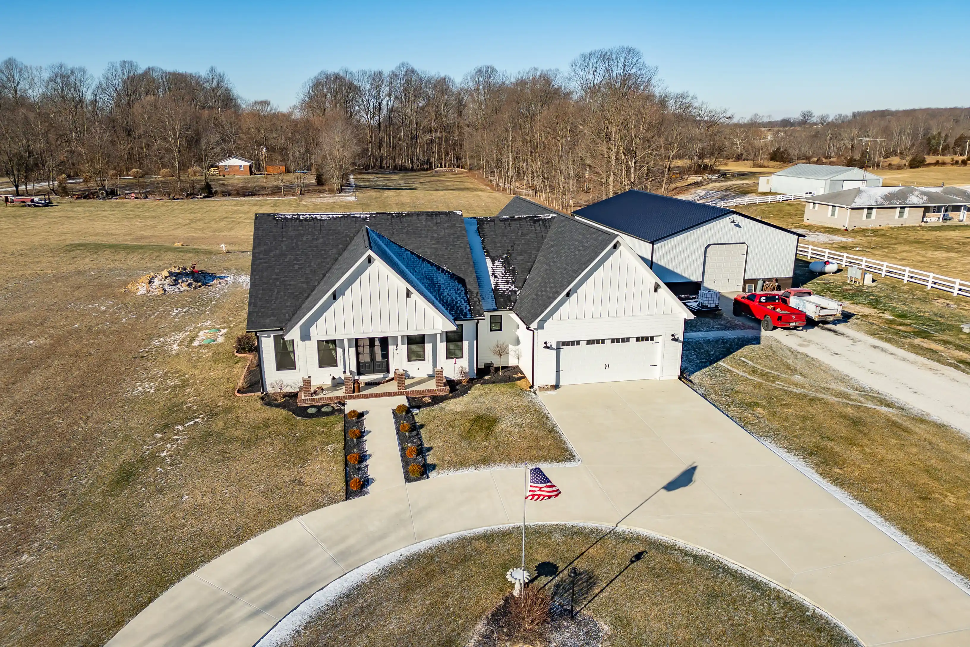 Aerial view of a modern farmhouse with white siding, black roof, and attached garage, next to a large metal outbuilding, with an American flag in the front yard and a red sports car parked nearby.