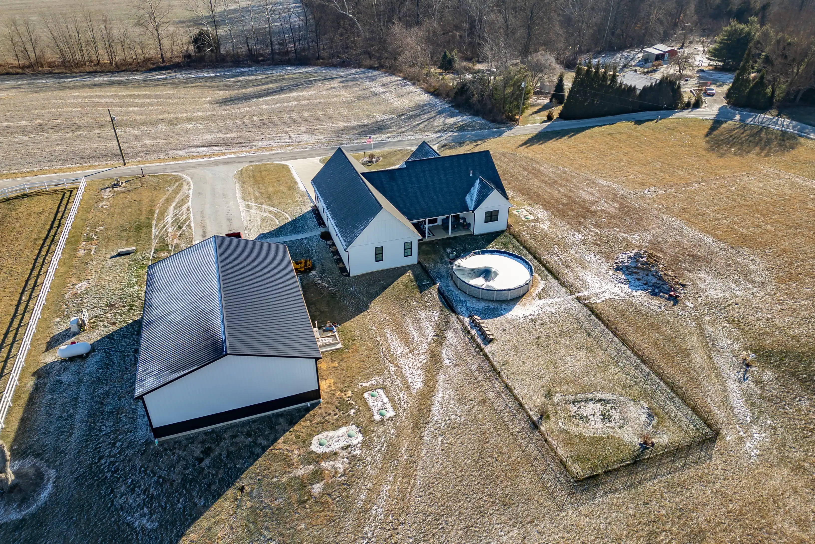 Aerial view of a rural residential property with a house, detached garage, above-ground pool, and surrounding fields in a sparse snowy landscape.
