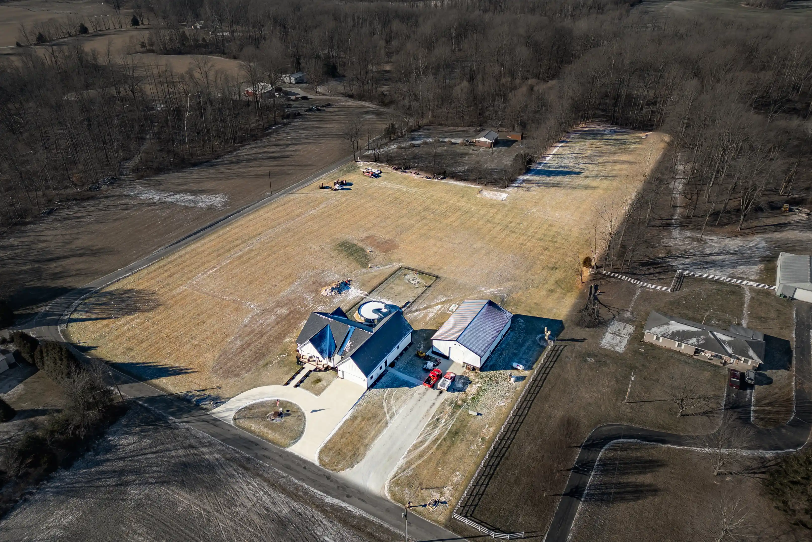 Aerial view of a rural property with a large blue-roofed house, adjacent buildings, a circular driveway, a swimming pool, and surrounding barren fields and trees.