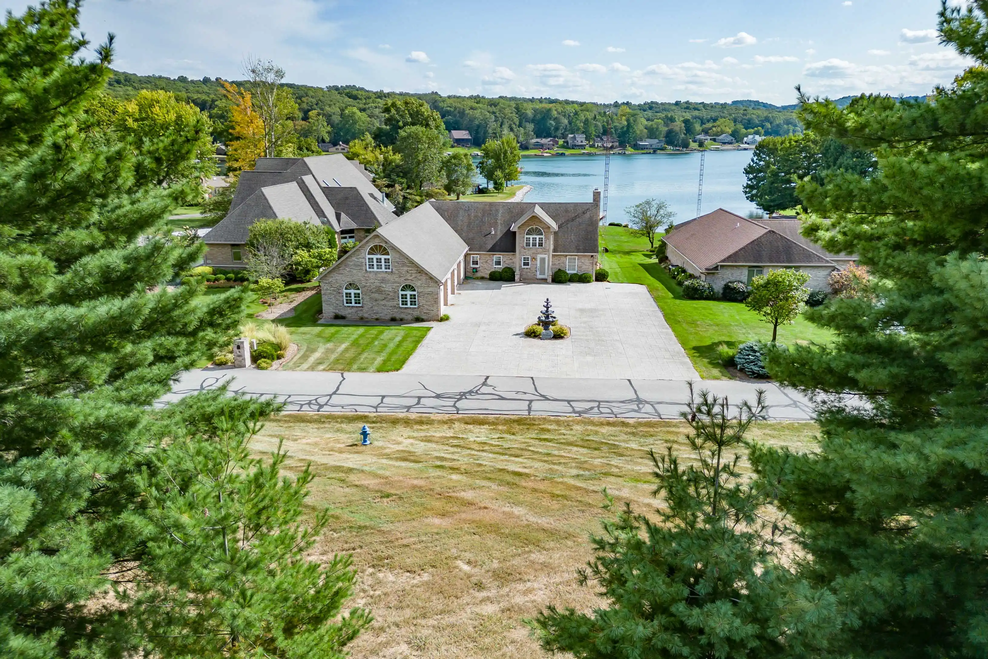 Aerial view of a spacious residential property with a large driveway, surrounded by trees, overlooking a lake with other houses in the distance.