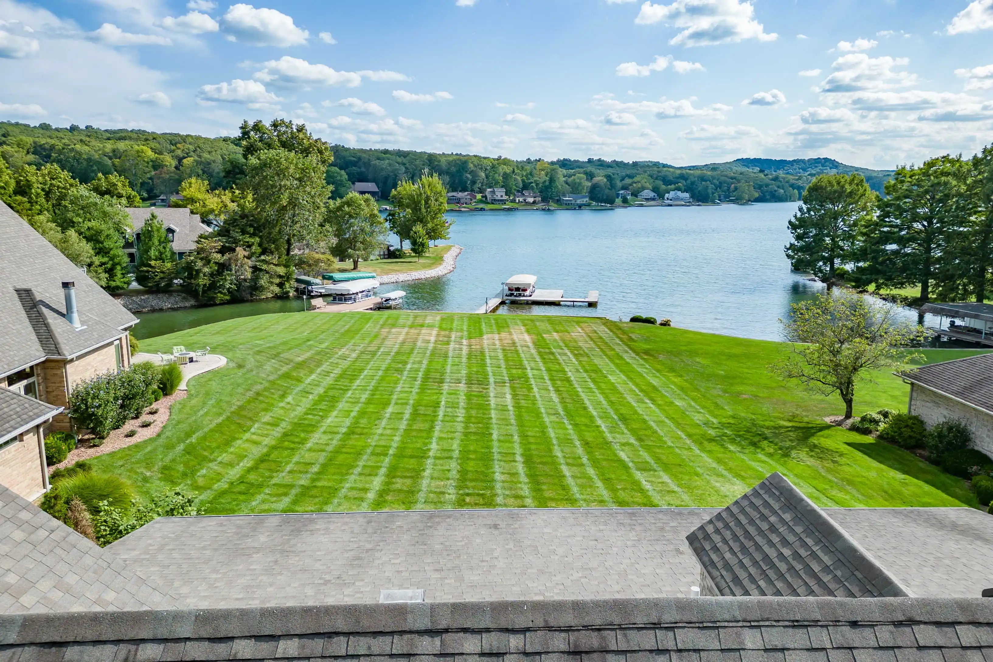 Aerial view of a well-manicured lawn leading to a lake with docks and boats, surrounded by residential homes and trees under a partly cloudy sky.