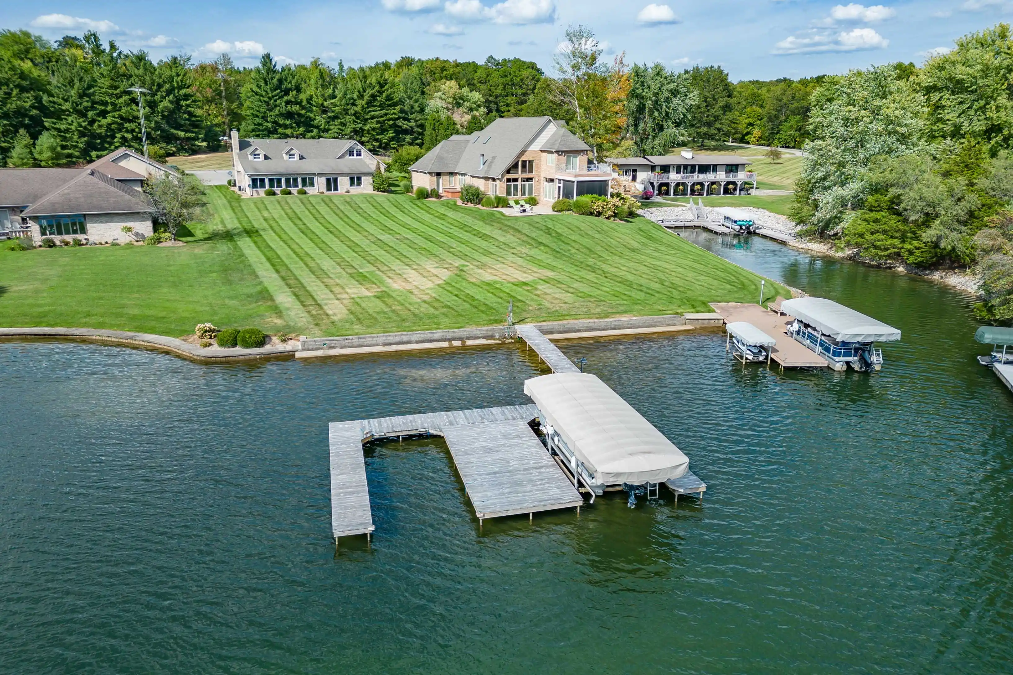 Aerial view of a large residential property with manicured lawn, multiple houses, and a private dock with covered boats on a calm body of water.