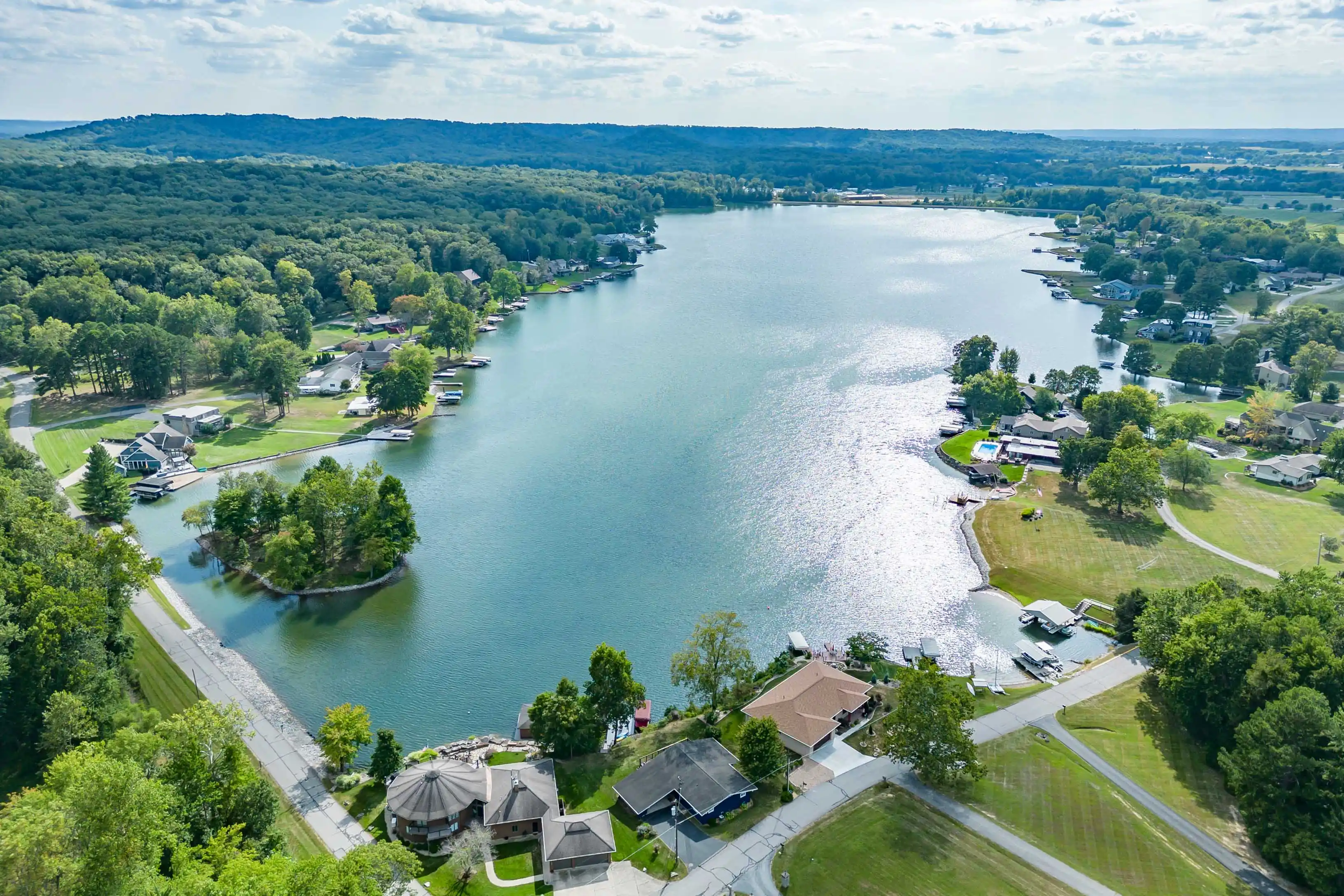 Aerial view of a serene lake surrounded by greenery with residential houses along the shoreline, a small island in the center, and distant hills under a cloudy sky.