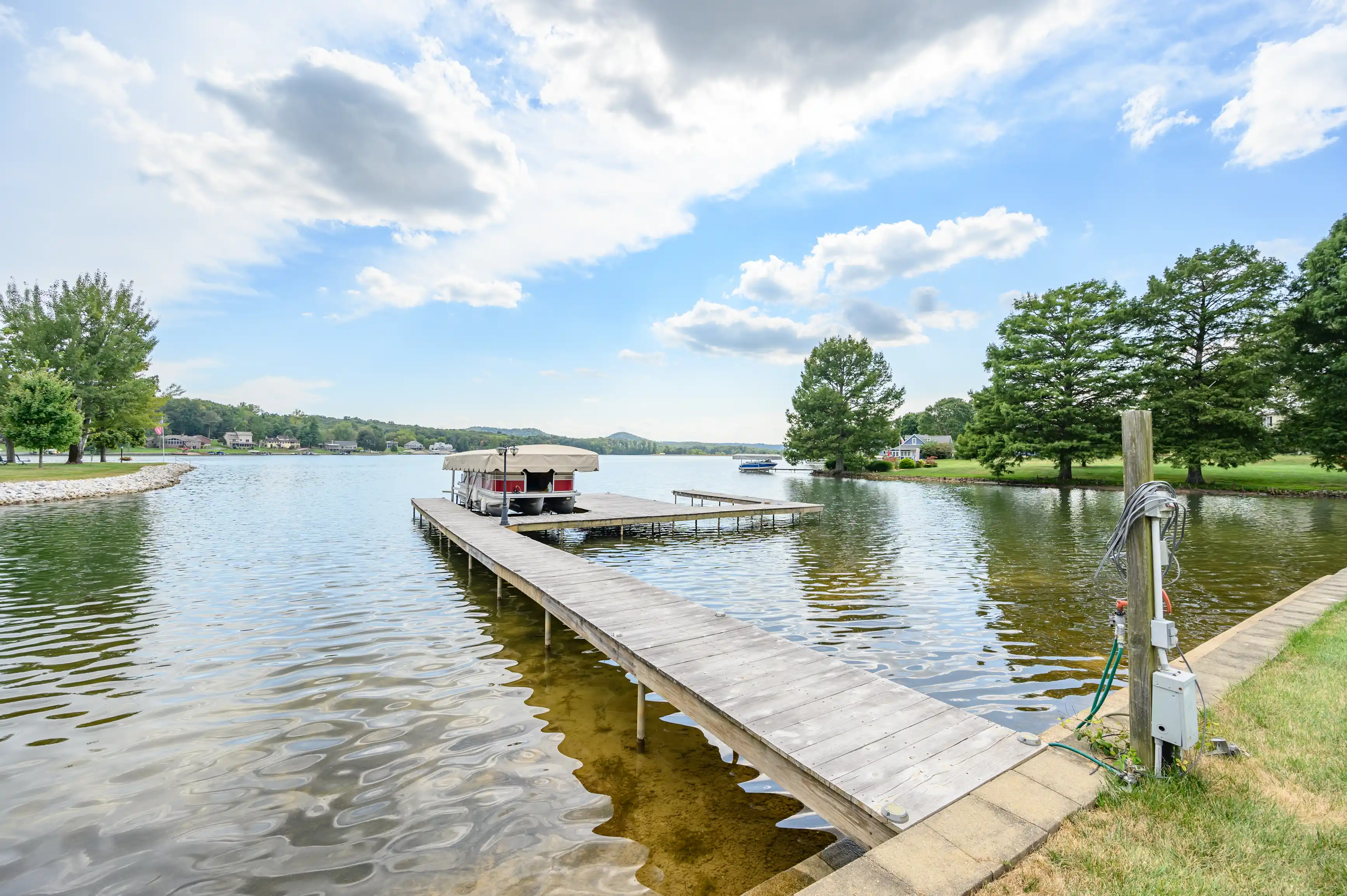 A tranquil lakeside scene with a wooden dock extending into the water, a covered pontoon boat moored at the end, surrounded by lush green trees under a partly cloudy sky.