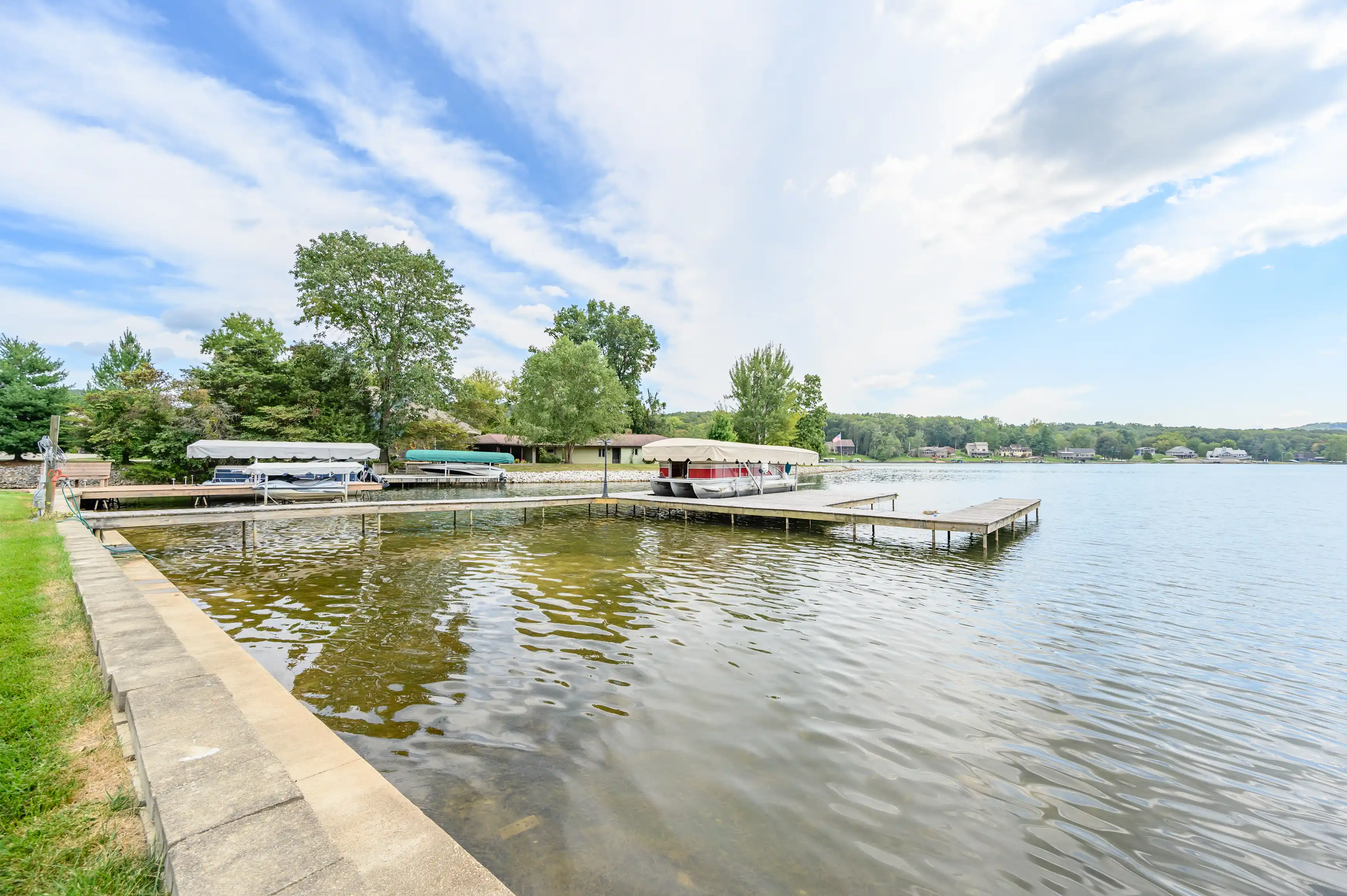 Scenic view of a calm lake with a dock, boats covered with tarps, and surrounding greenery on a clear day.