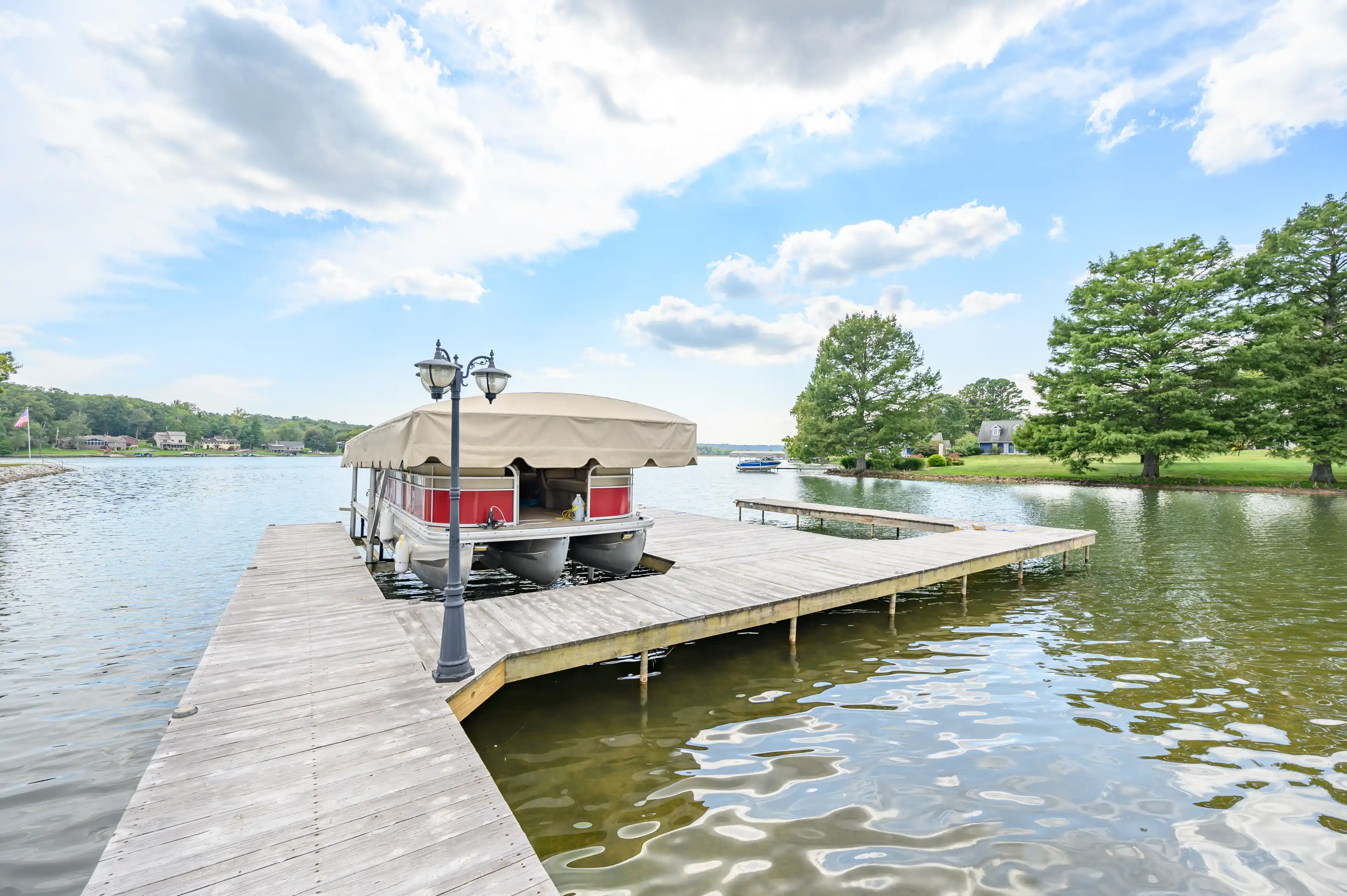 A pontoon boat moored at a wooden dock on a peaceful lake with greenery and a blue sky with clouds.