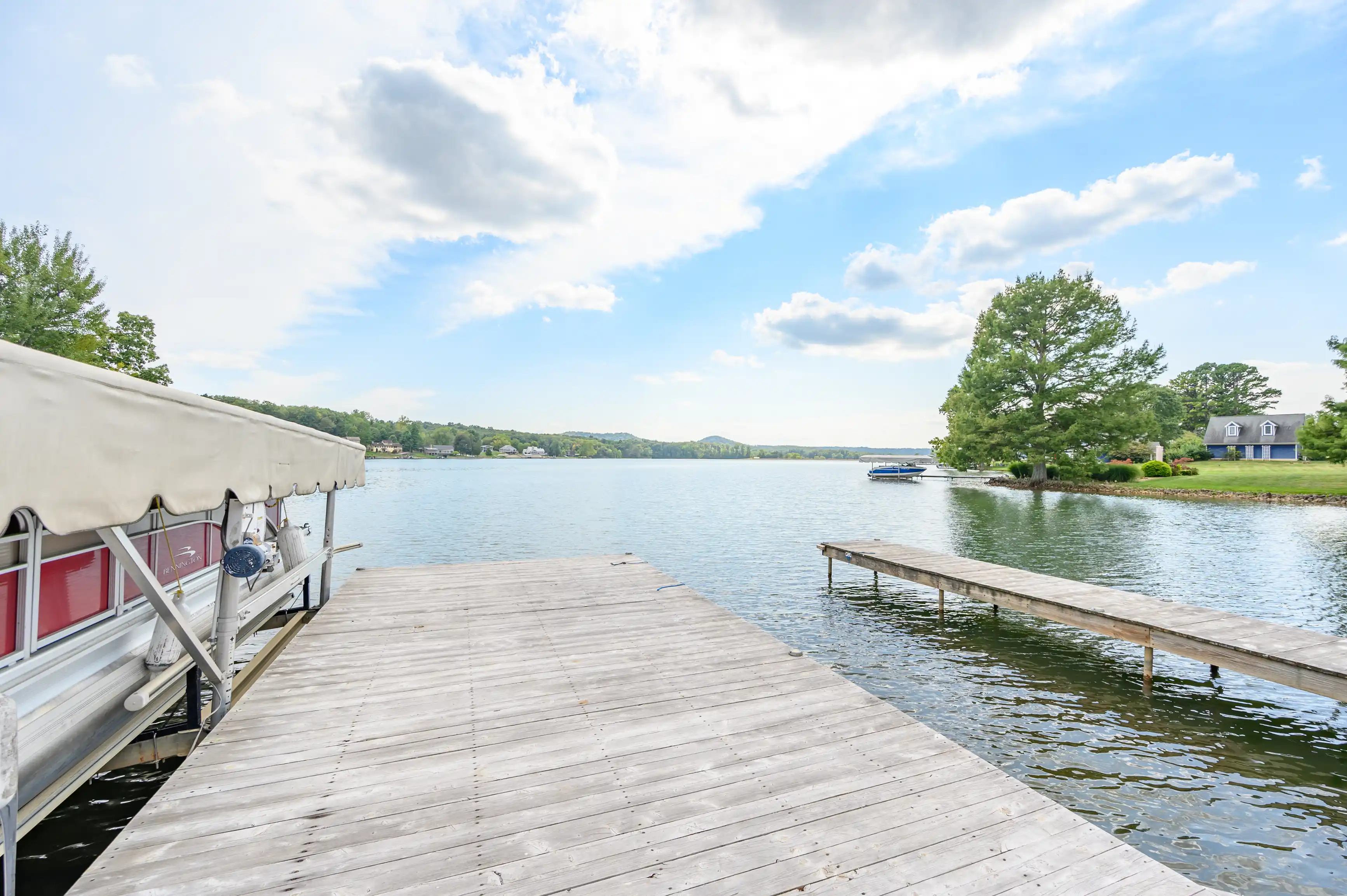 A serene lakeside view with a wooden dock extending into the water, a covered pontoon boat on the left, and a landscape with houses and trees in the background under a blue sky with clouds.
