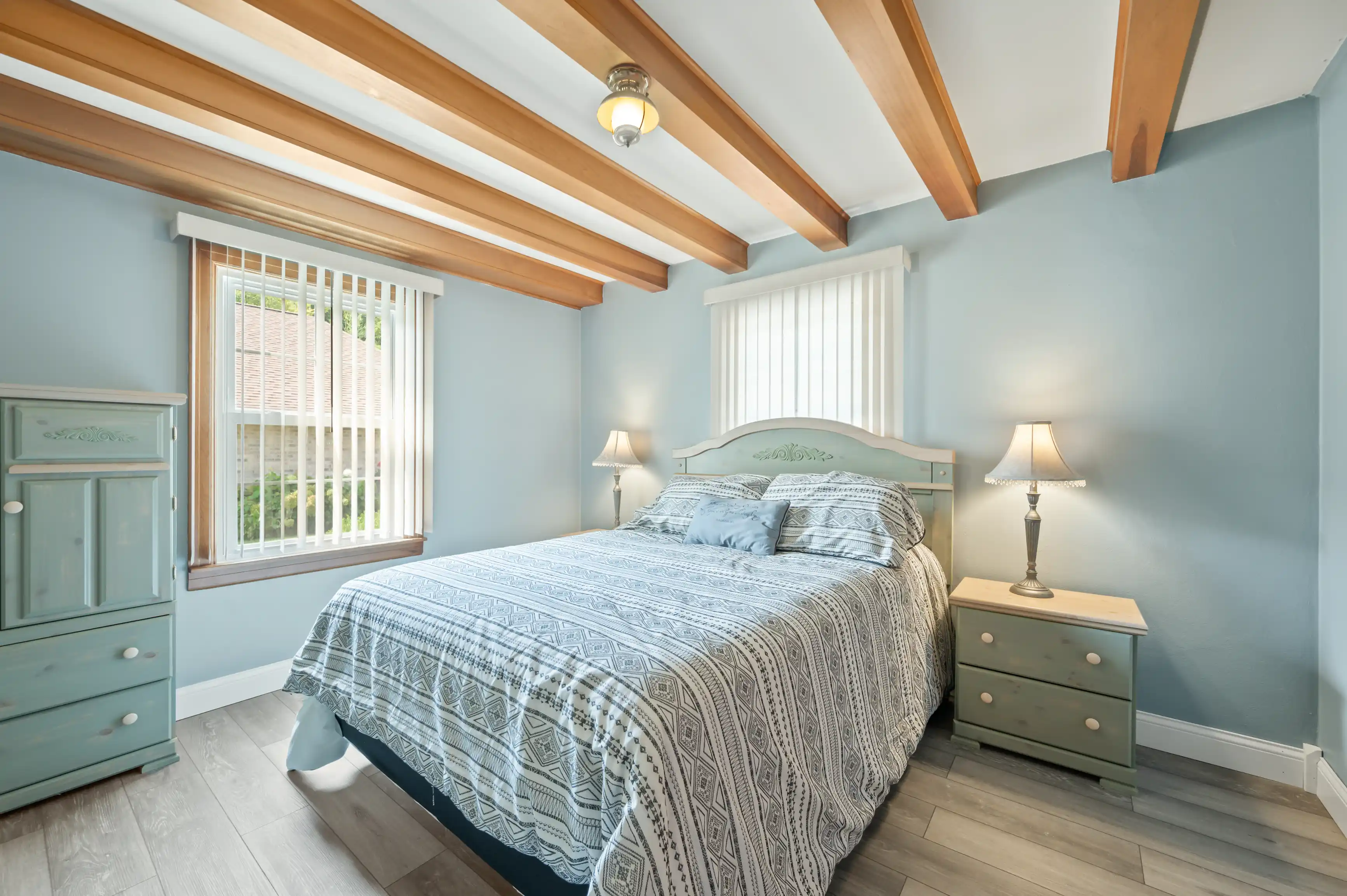 A cozy bedroom featuring a queen-sized bed with patterned bedding, matching pale green wooden furniture, light blue walls with white trims, exposed wooden beams, and a large window with vertical blinds.