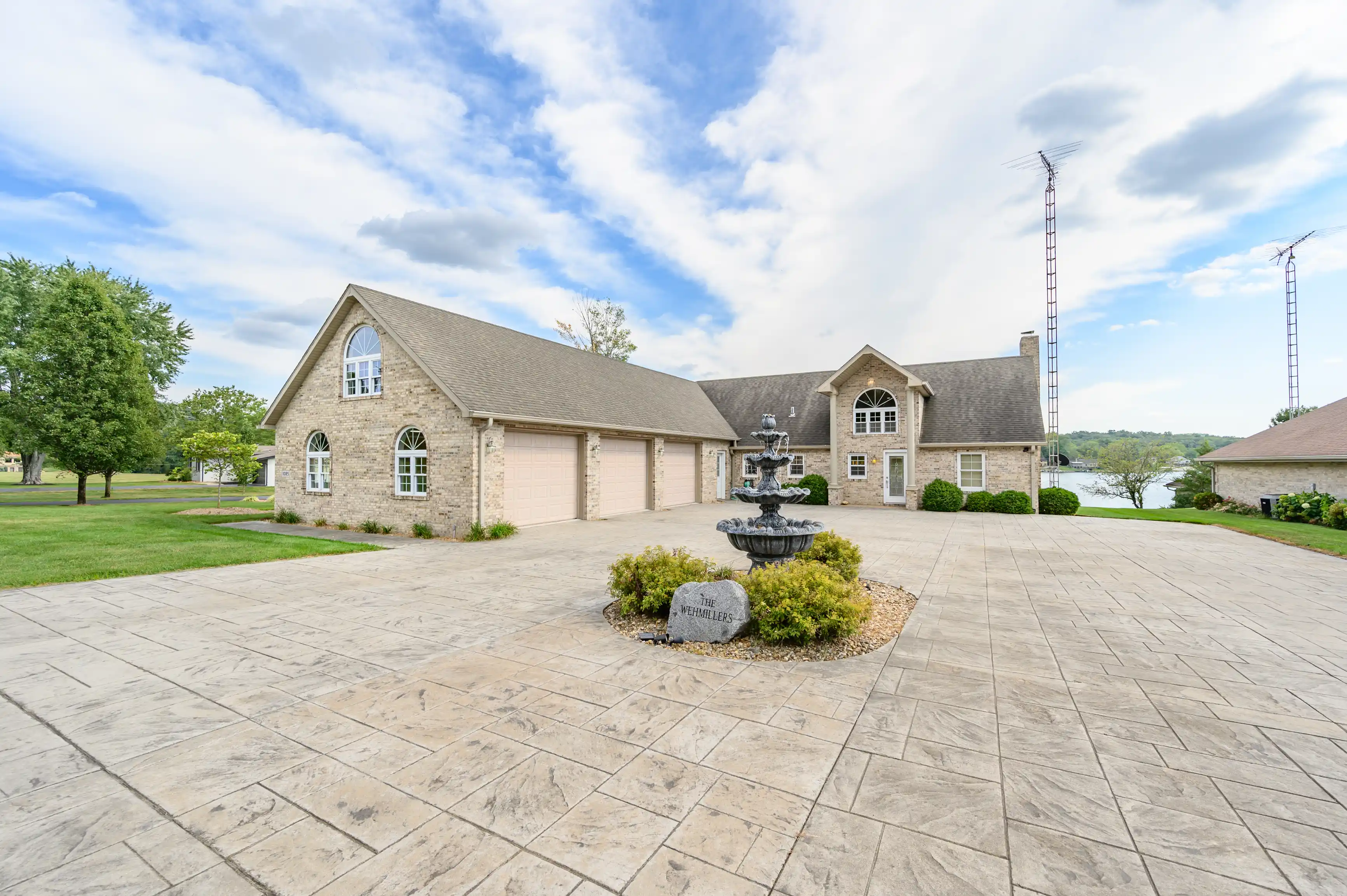 Spacious single-family home with stone facade and attached three-car garage, featuring a large paved driveway and central fountain in a landscaped front yard, set against a partly cloudy sky with antennas in the background and a view of a water body behind the property.