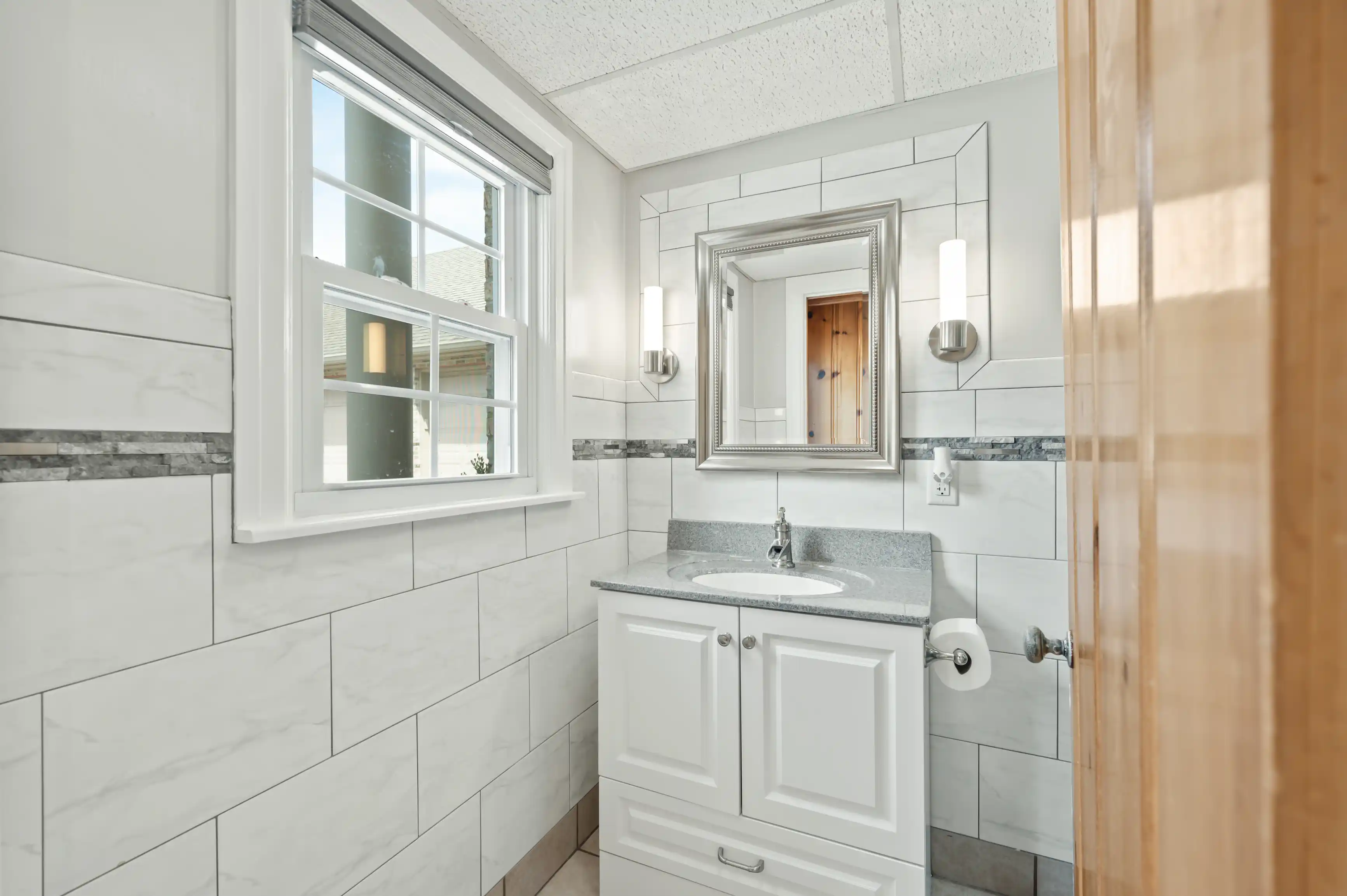 Bright bathroom interior with a large window, subway tiled walls, a vanity with a mounted sink and mirror, and modern wall sconces.