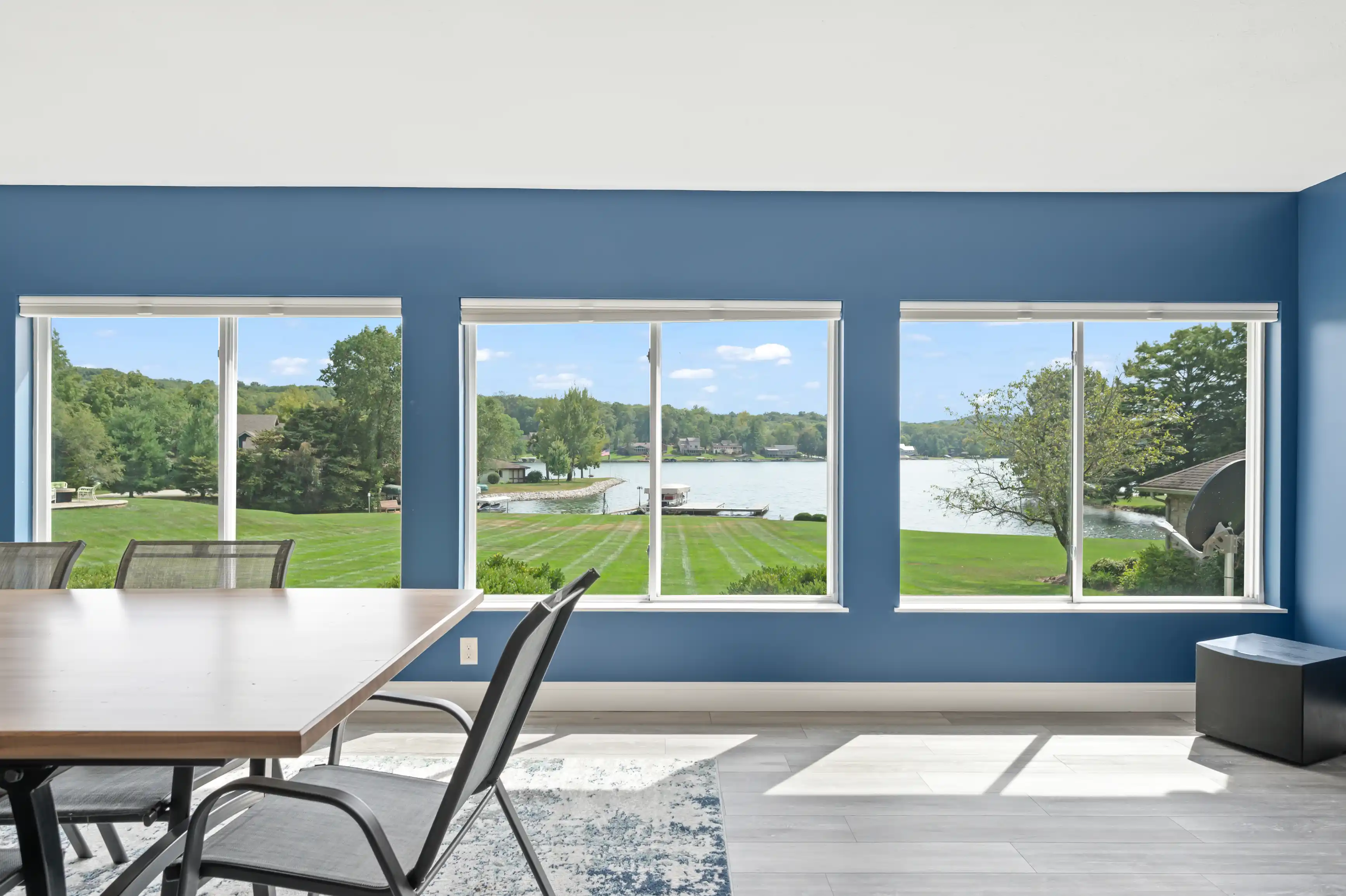 Spacious room with large windows overlooking a lake with green lawn and dock, featuring a minimalist dining table and blue accent wall.
