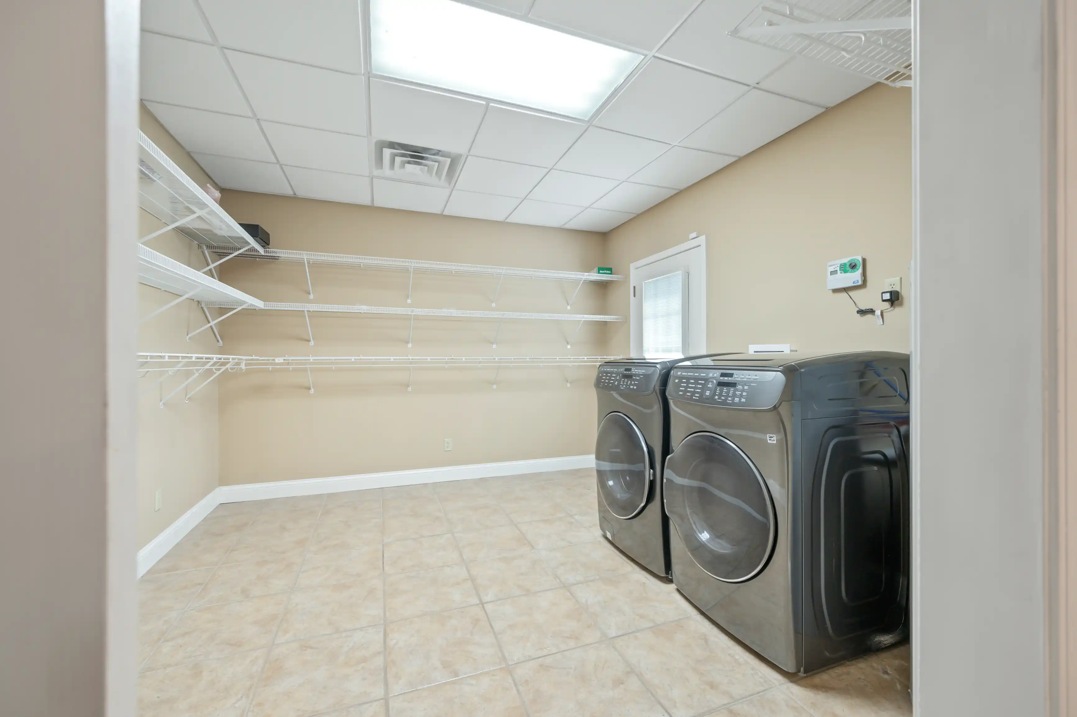 A spacious laundry room with beige walls, tiled flooring, a set of modern washing machine and dryer, and multiple white wire shelves.