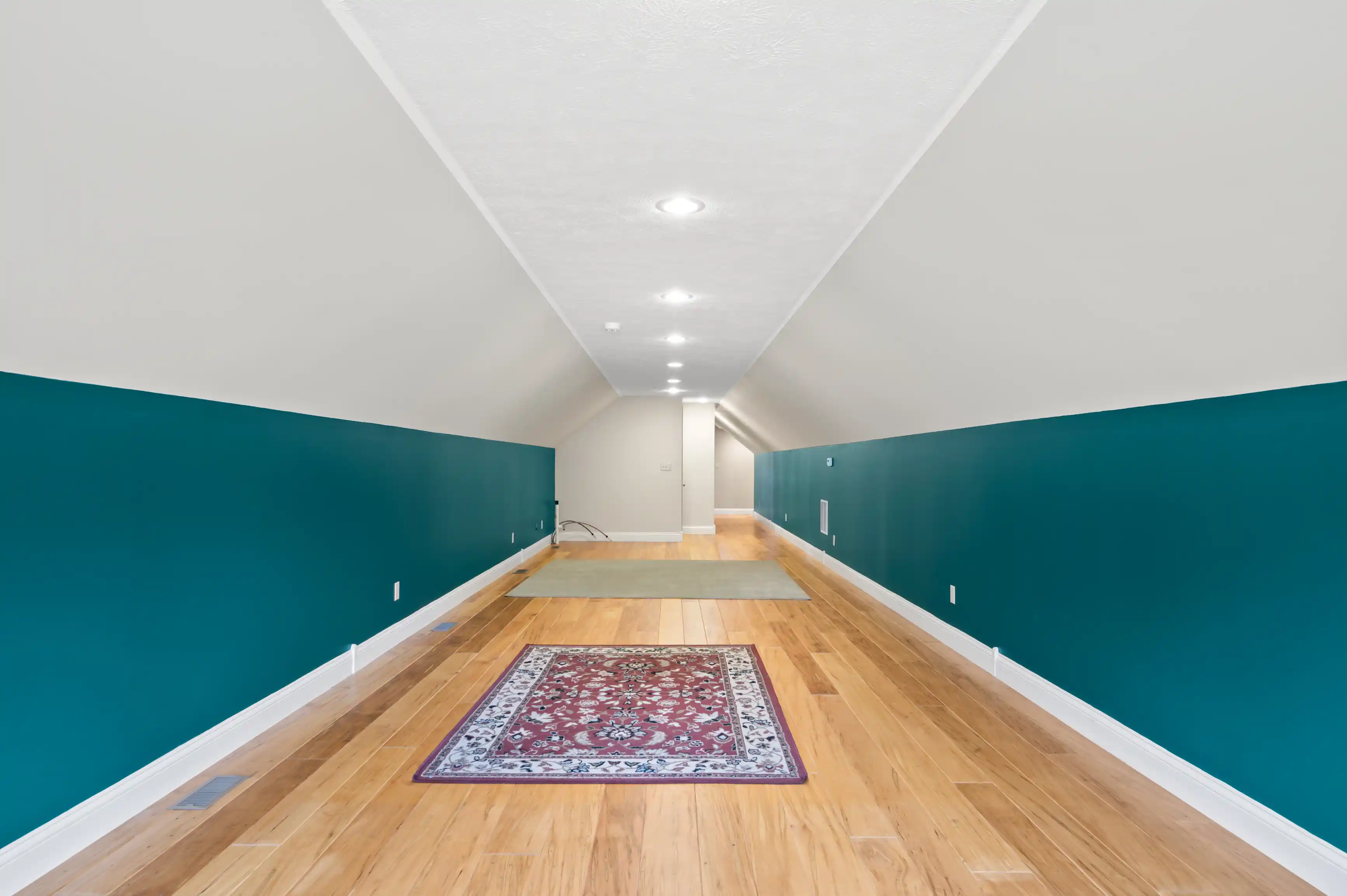 A long, narrow hallway with hardwood floors, white ceiling, and dark teal lower walls, featuring recessed lighting and a decorative red and blue rug.