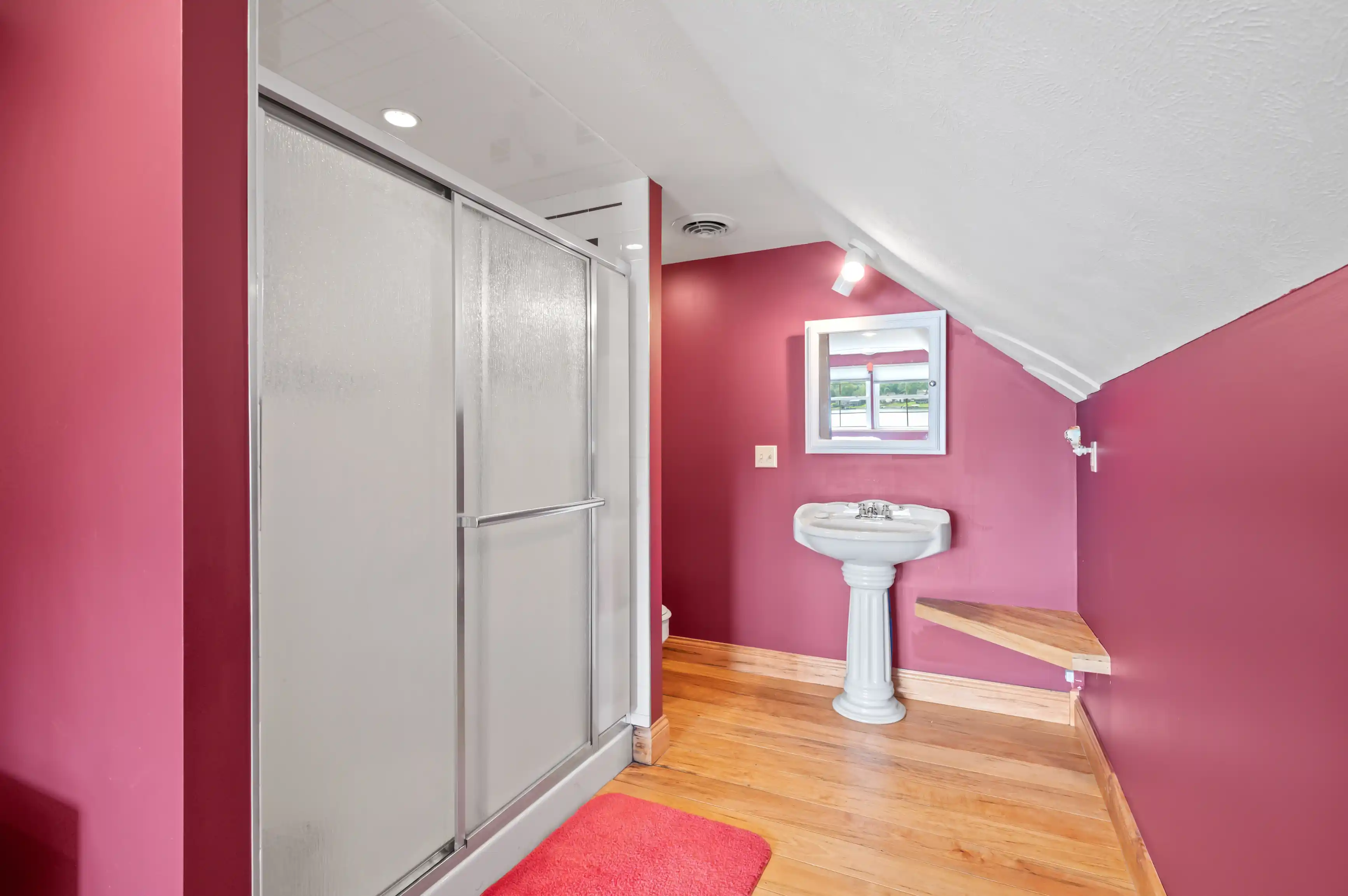 Interior of a bathroom with magenta walls, hardwood floors, a glass shower door, pedestal sink, and a small wooden shelf under a window with a view outside.