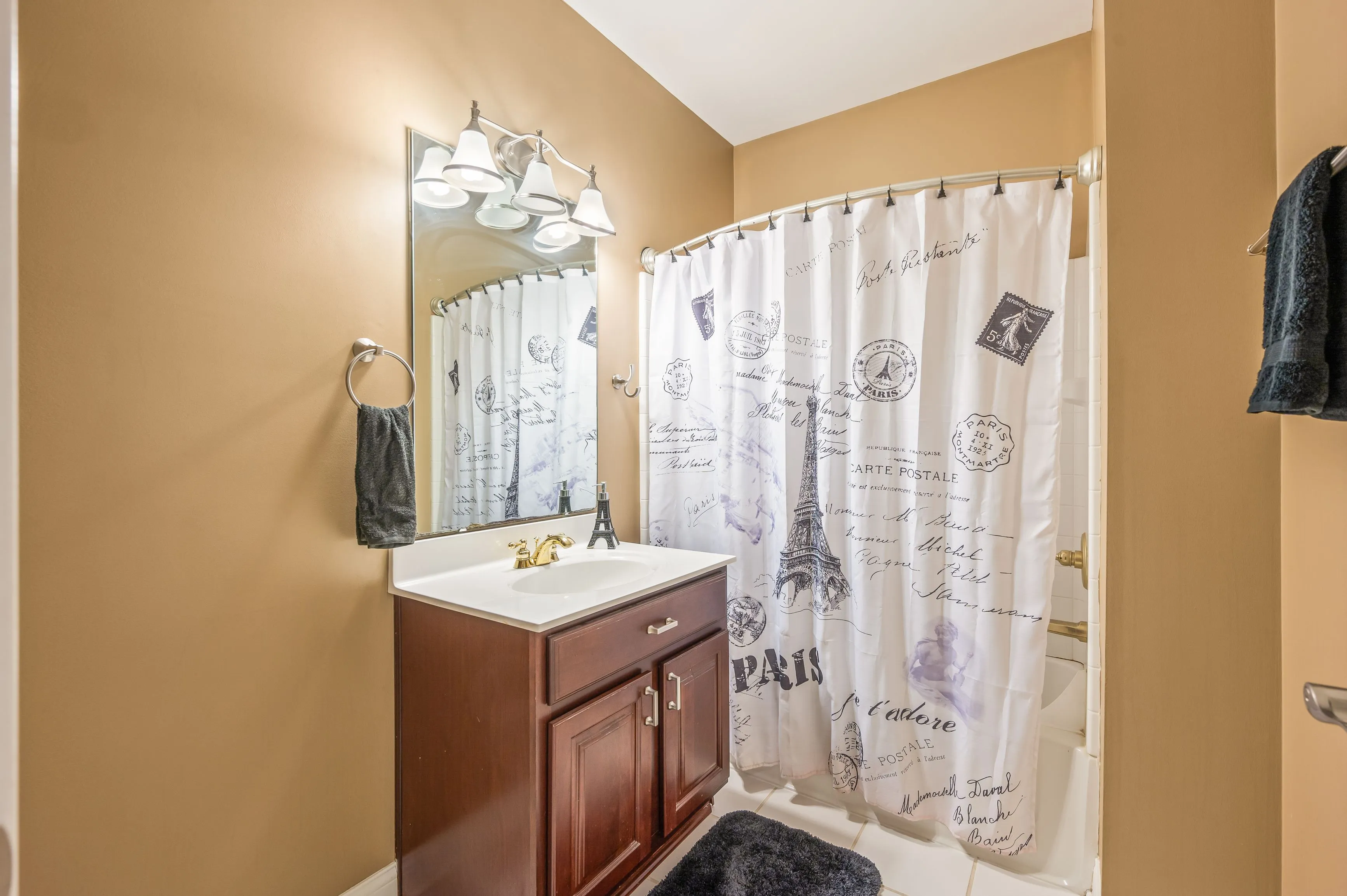 Compact bathroom with beige walls, shower curtain with Paris landmarks, wooden vanity cabinet and wall-mounted towel.