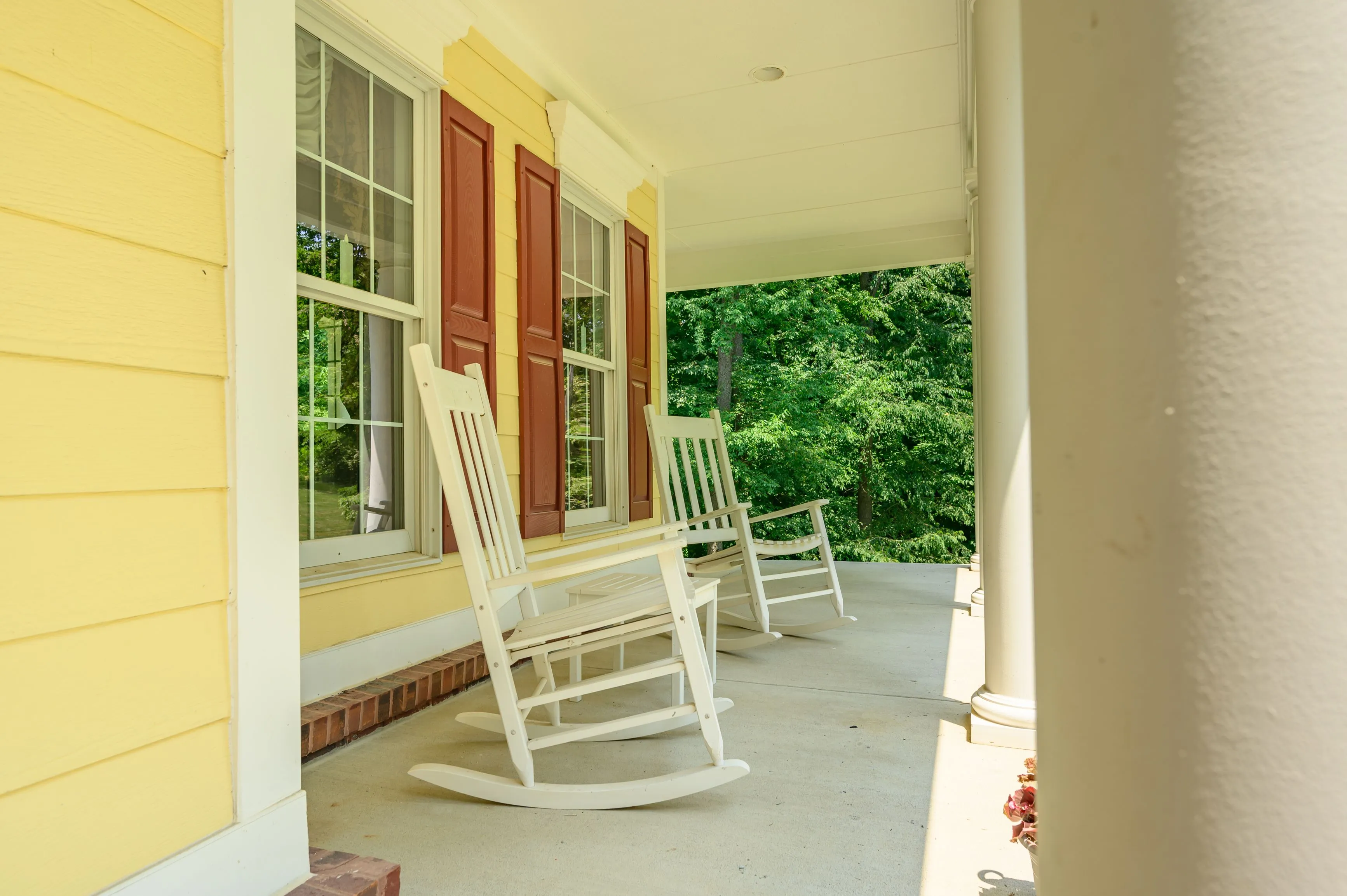  A sunny porch with two white rocking chairs, red shutters on the windows, and a view of green bushes and trees.