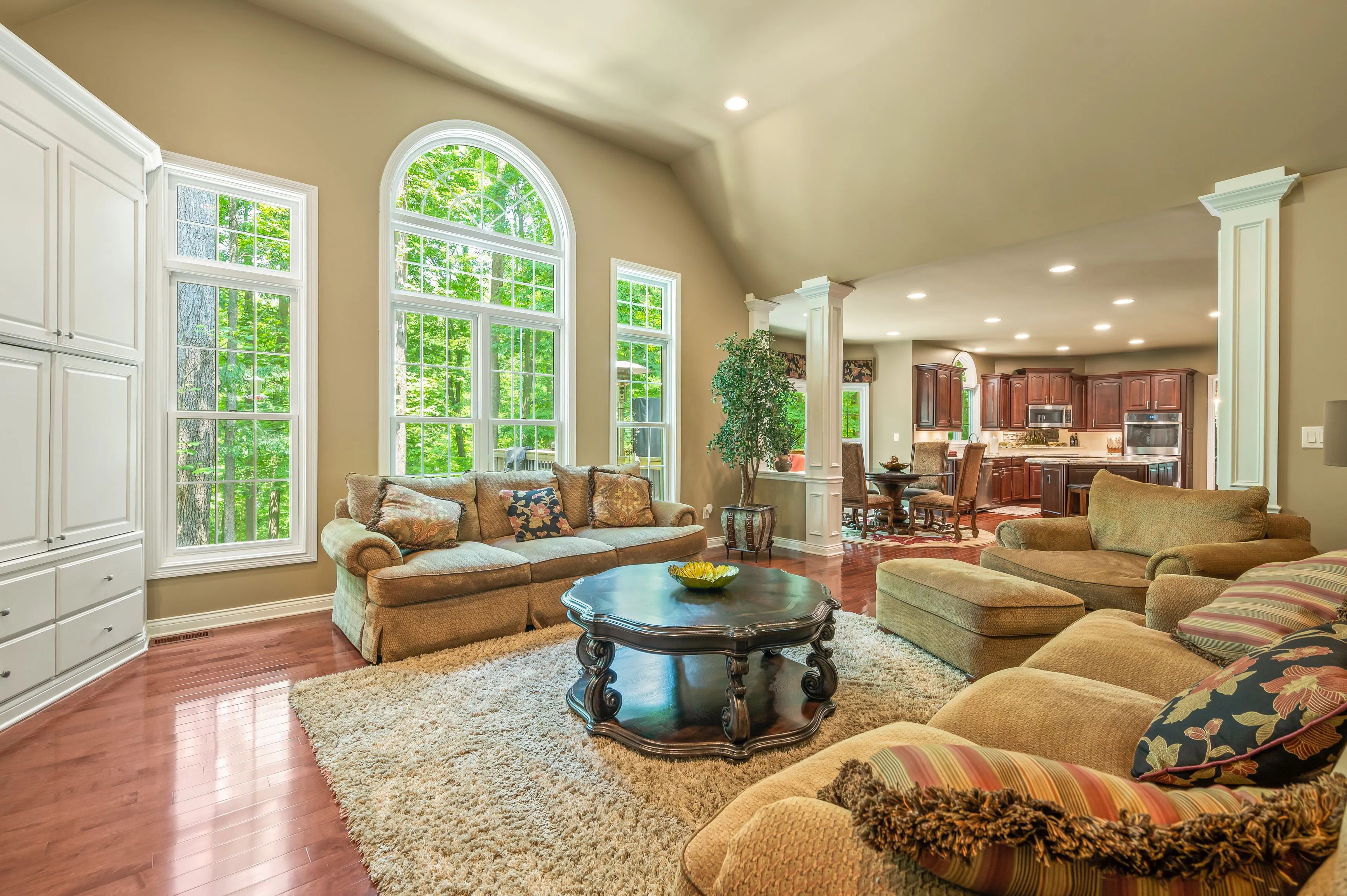 Elegant living room with large arched windows, tan sofas, a round coffee table, and a view into an adjoining kitchen area.