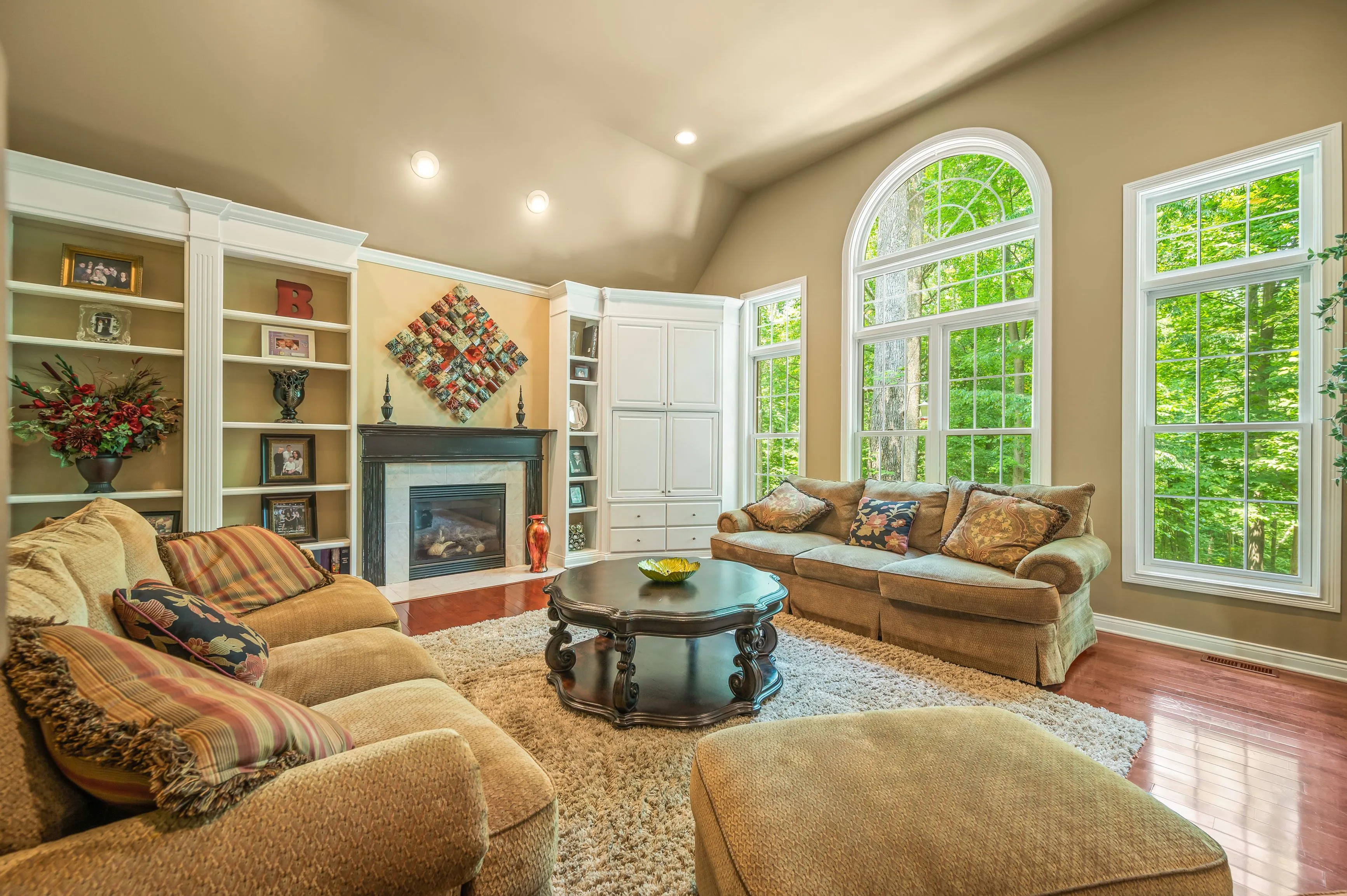 Bright and cozy living room interior with plush sofas, a fireplace, built-in bookshelves, large arched windows with a view of greenery, hardwood floors, and a central coffee table.