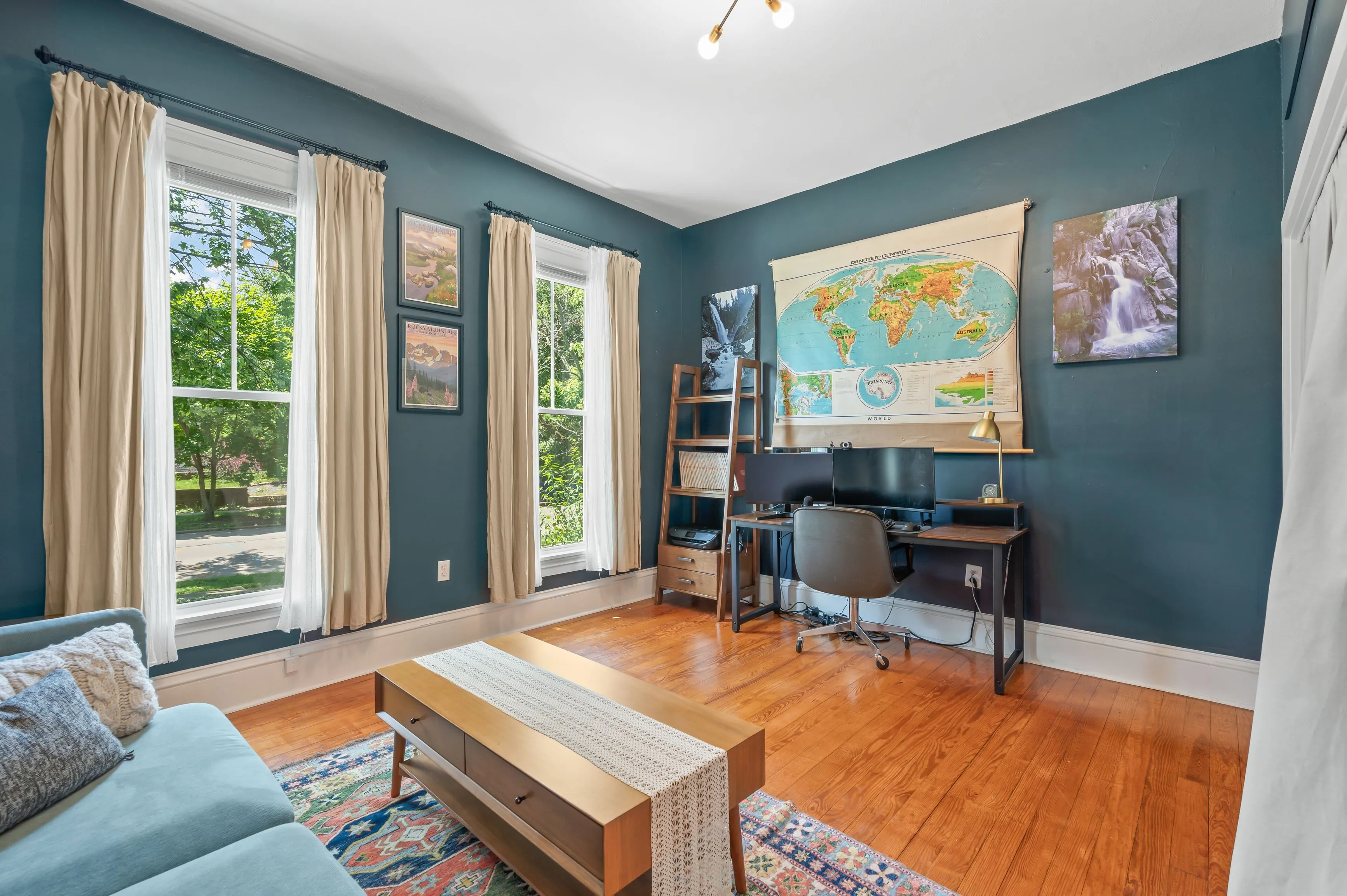 Cozy living room with hardwood floors, a blue sofa, wooden coffee table, and a world map hanging above a computer desk by the window.