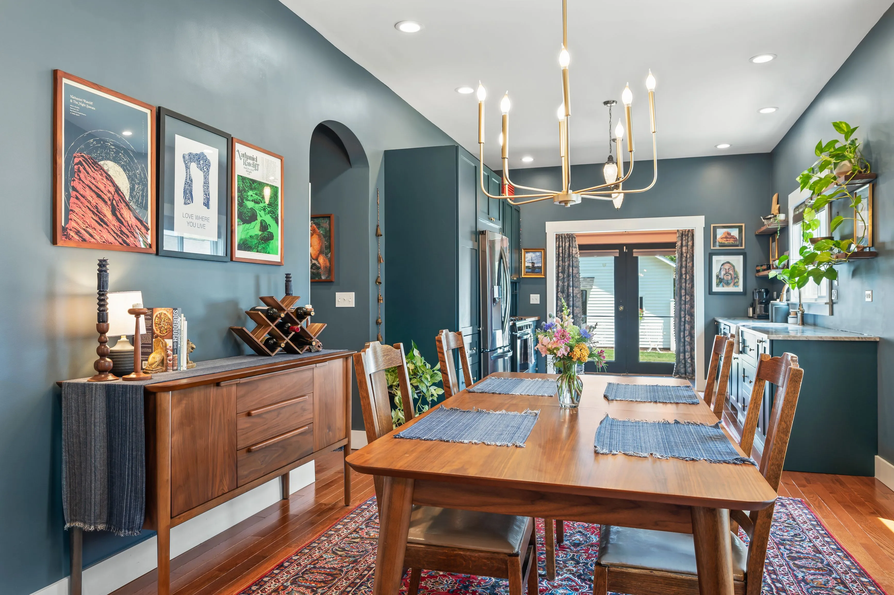 Elegantly decorated dining room with a wooden table, blue upholstered chairs, and a modern chandelier, surrounded by artwork on blue walls and a wooden sideboard on one side.