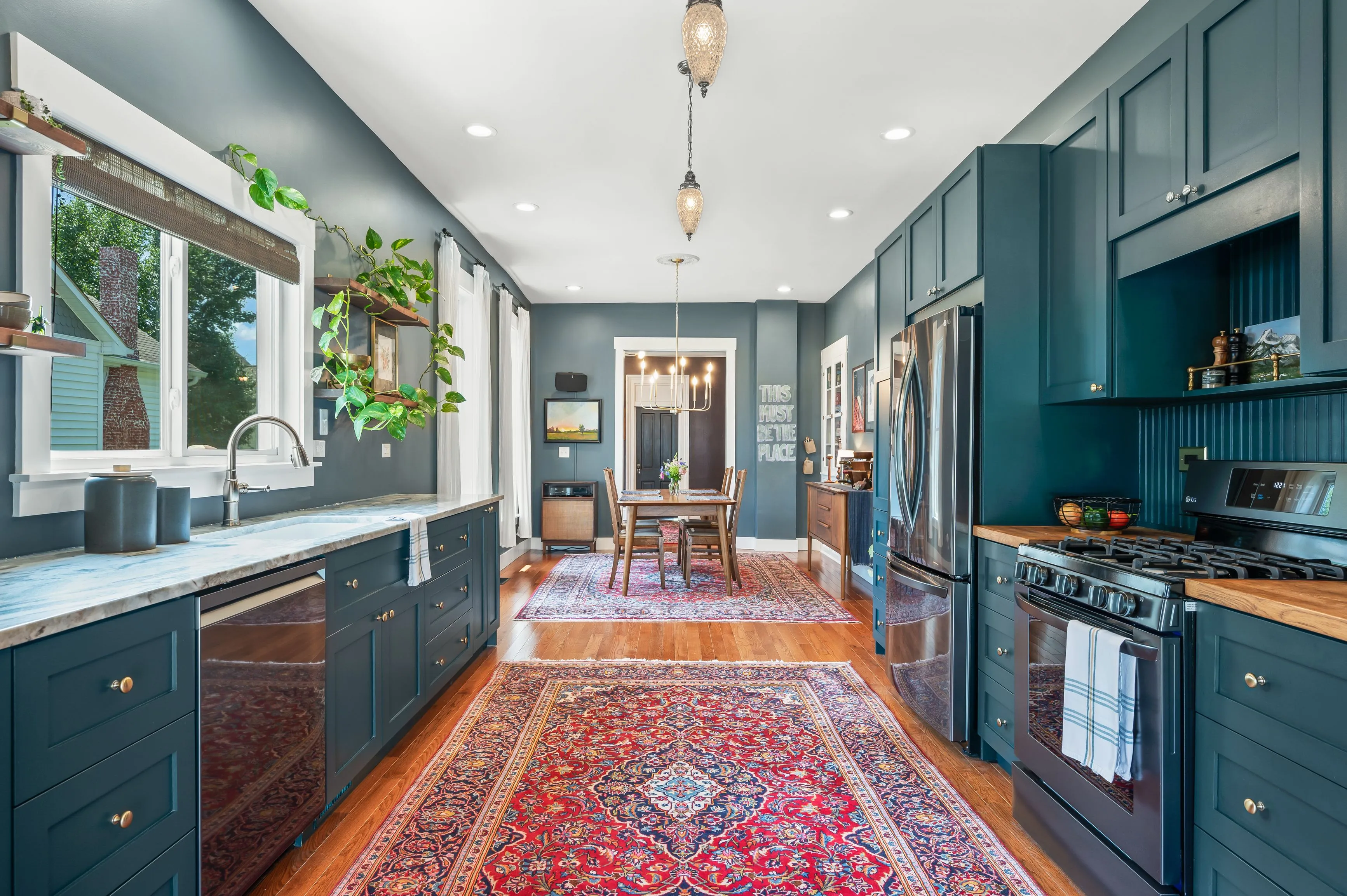 Elegant kitchen interior with dark blue cabinetry, stainless steel appliances, a colorful rug on hardwood floor, and pendant lighting, leading to a dining area.