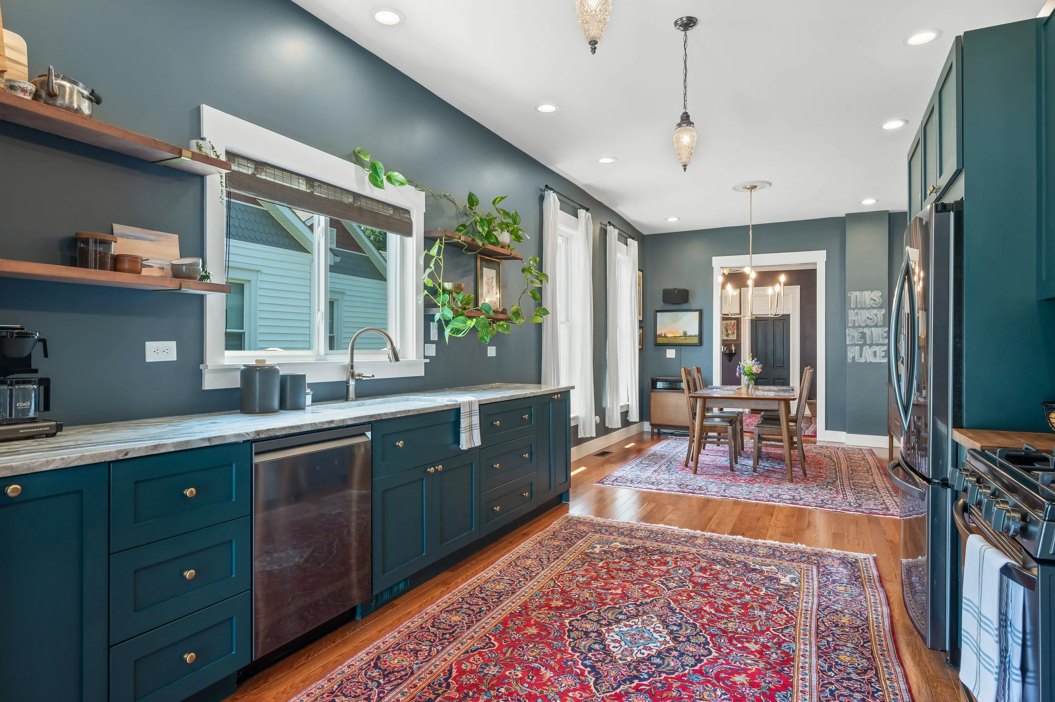 Elegant interior of a spacious kitchen with dark green cabinetry, stainless steel appliances, Persian rug on the hardwood floor, leading to a well-lit dining area.