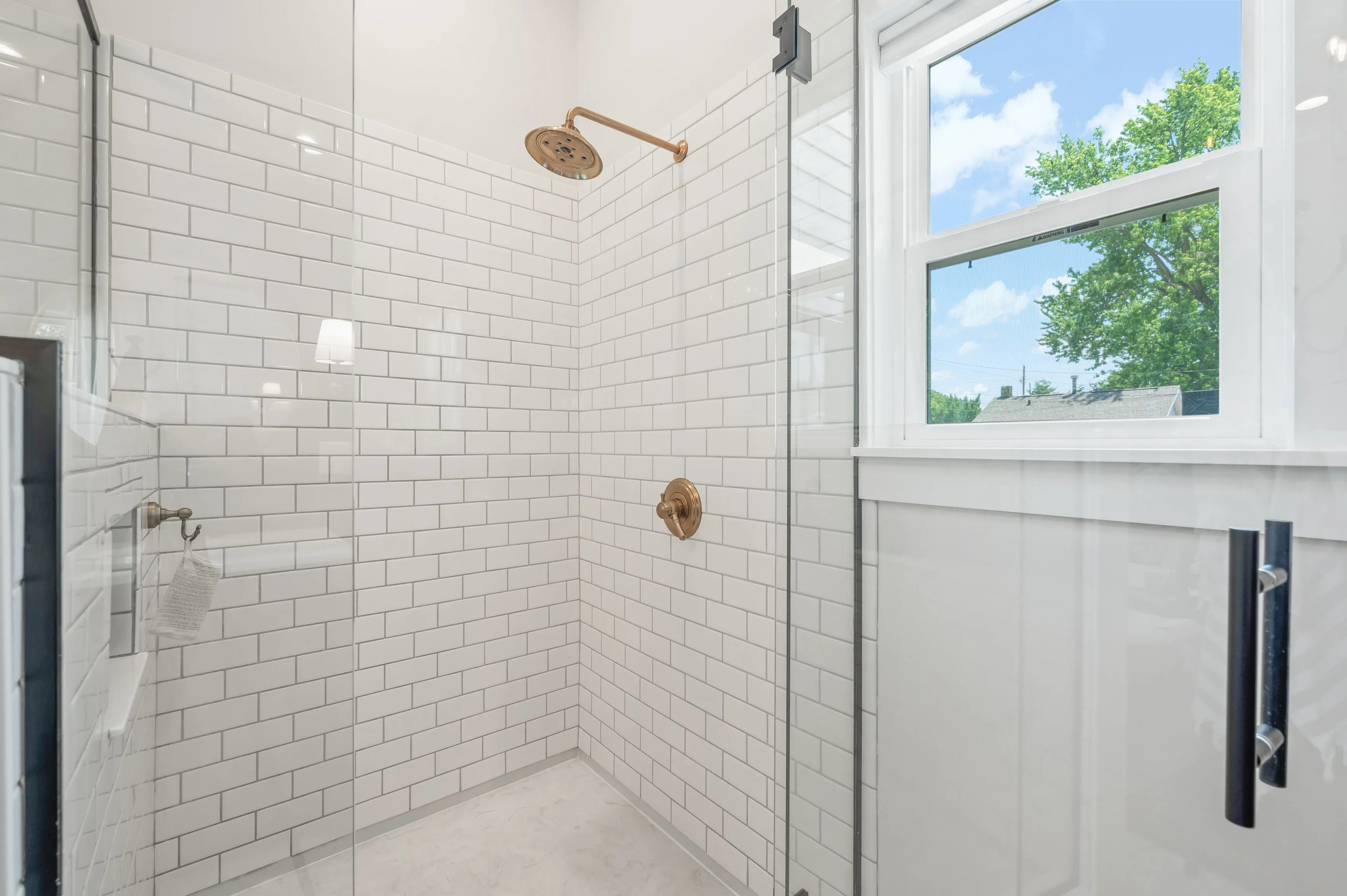 Modern bathroom with white subway tiles, glass shower door, and a window with a view of greenery outside.