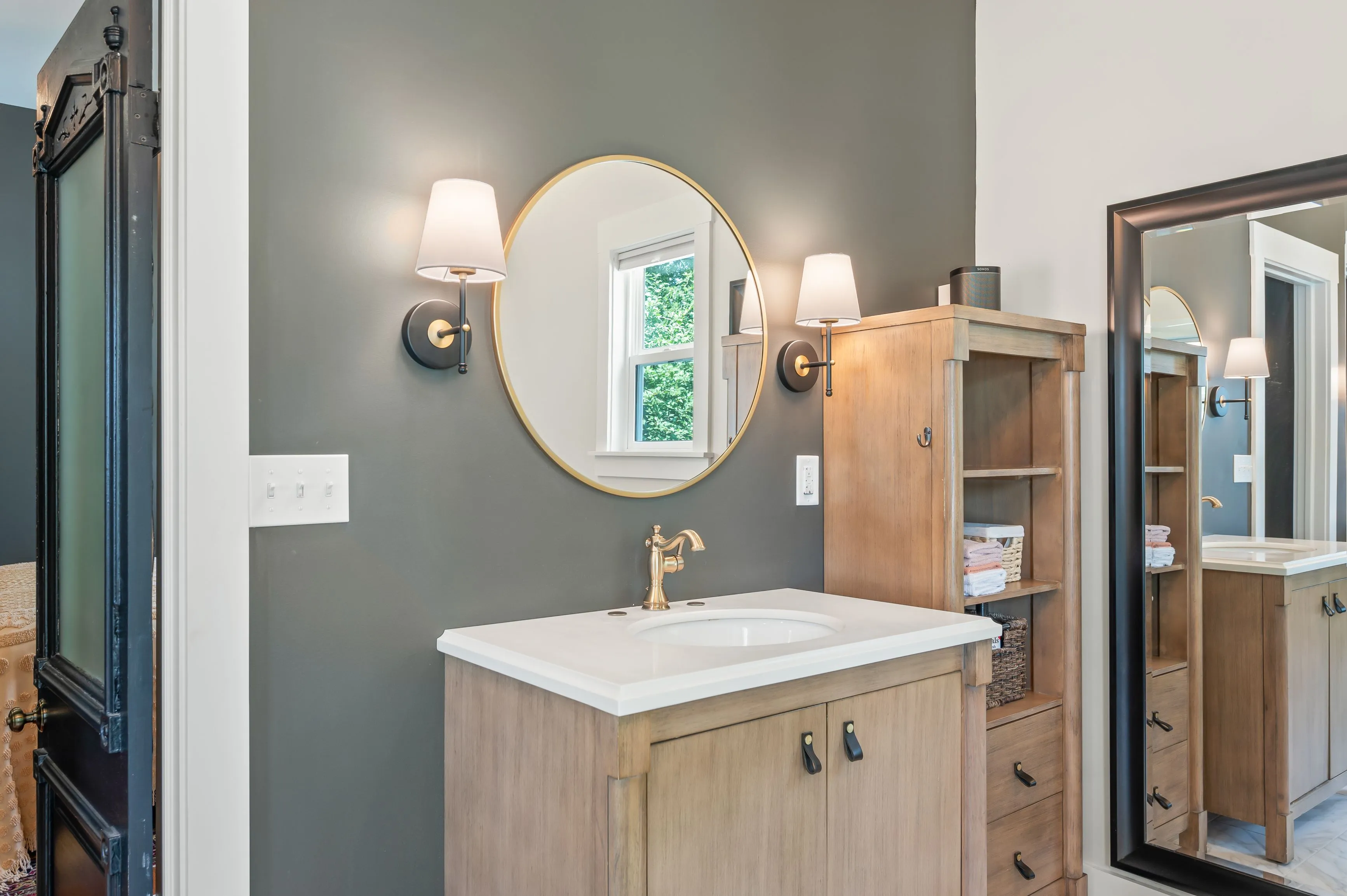 Modern bathroom with a gray wall, wooden vanity with a white sink, round mirror, and sconce lighting fixtures.