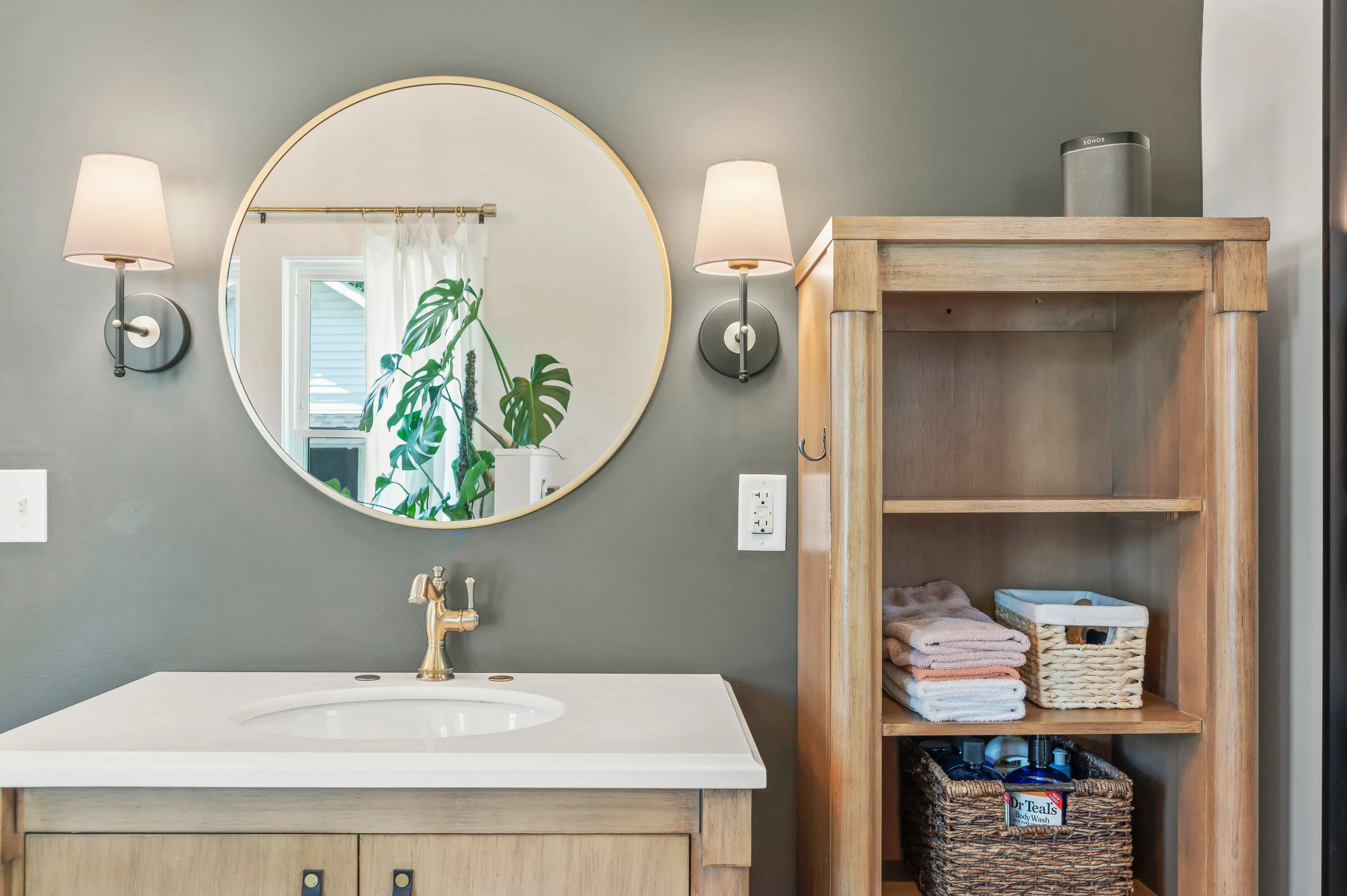 Modern bathroom interior with round mirror, wall sconces, and wooden shelving unit with towels.