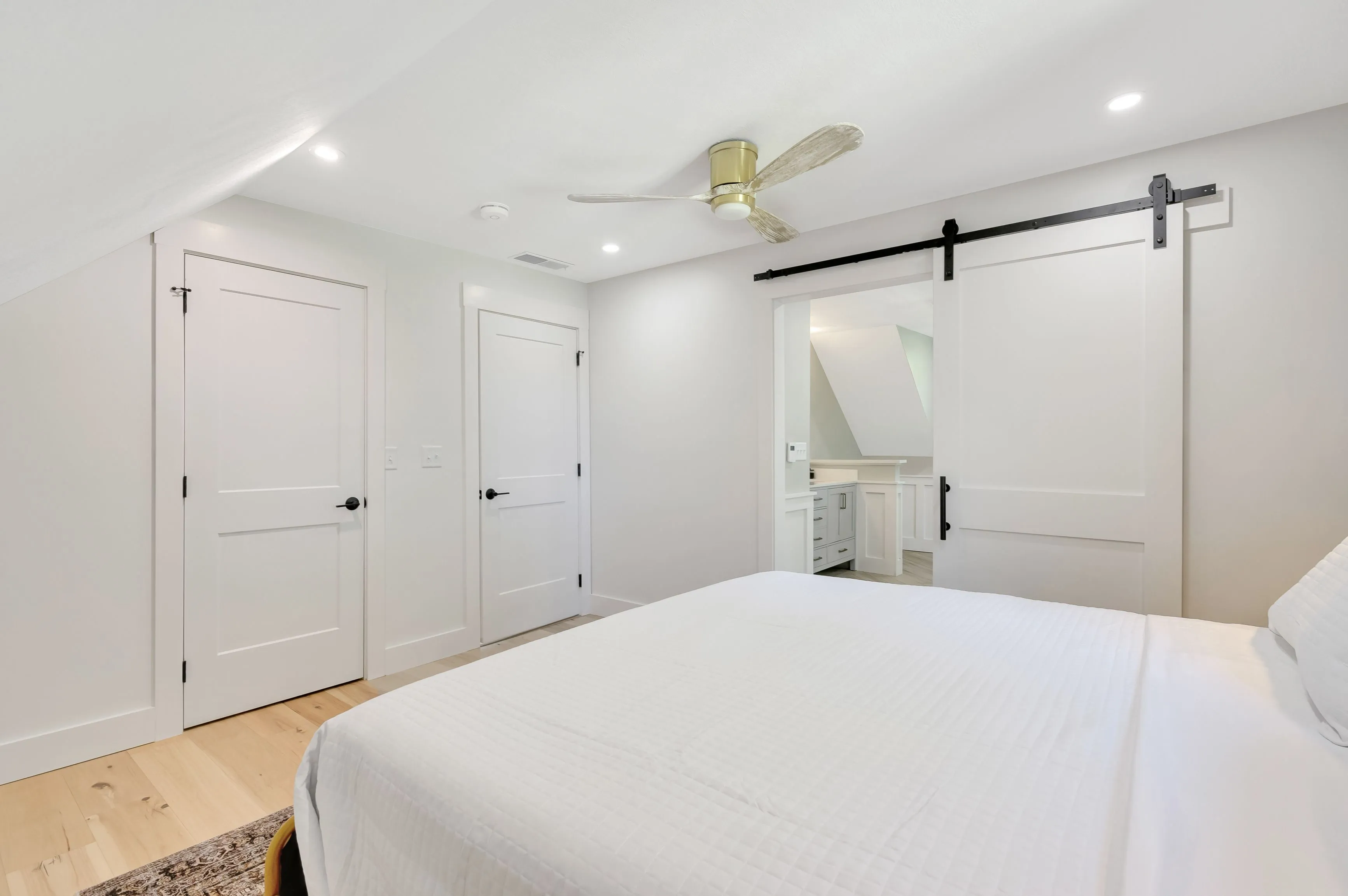 Bright contemporary bedroom with a large bed, white bedding, light wooden flooring, white walls, a ceiling fan, and a sliding barn door leading to an ensuite bathroom.