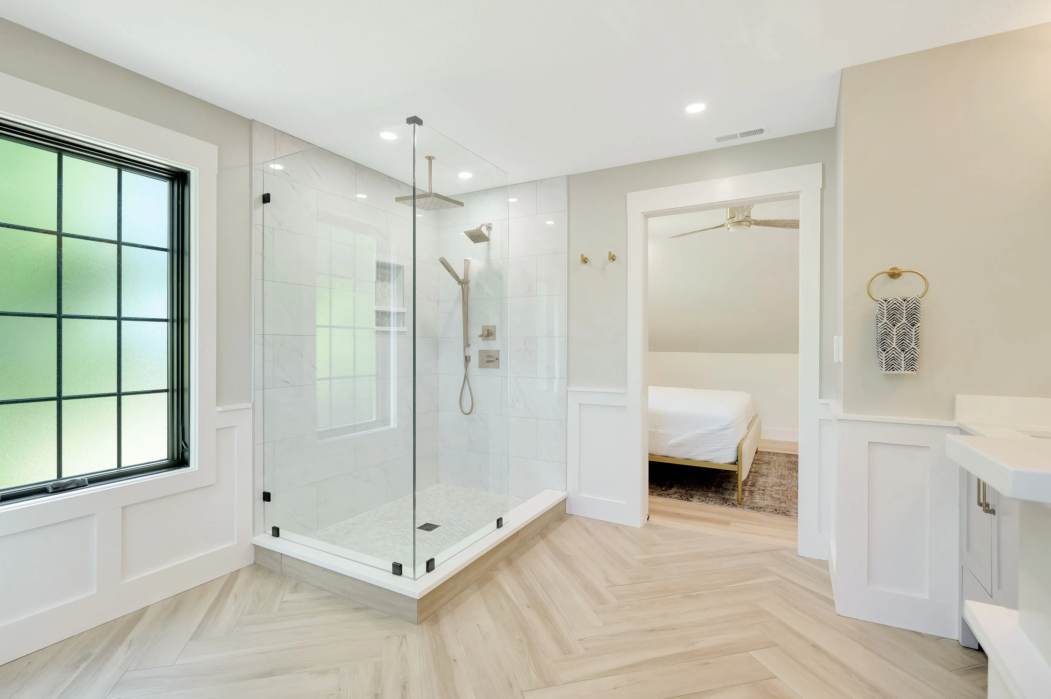 Modern bathroom interior with herringbone pattern floor, walk-in shower, double vanity, and view into bedroom area.