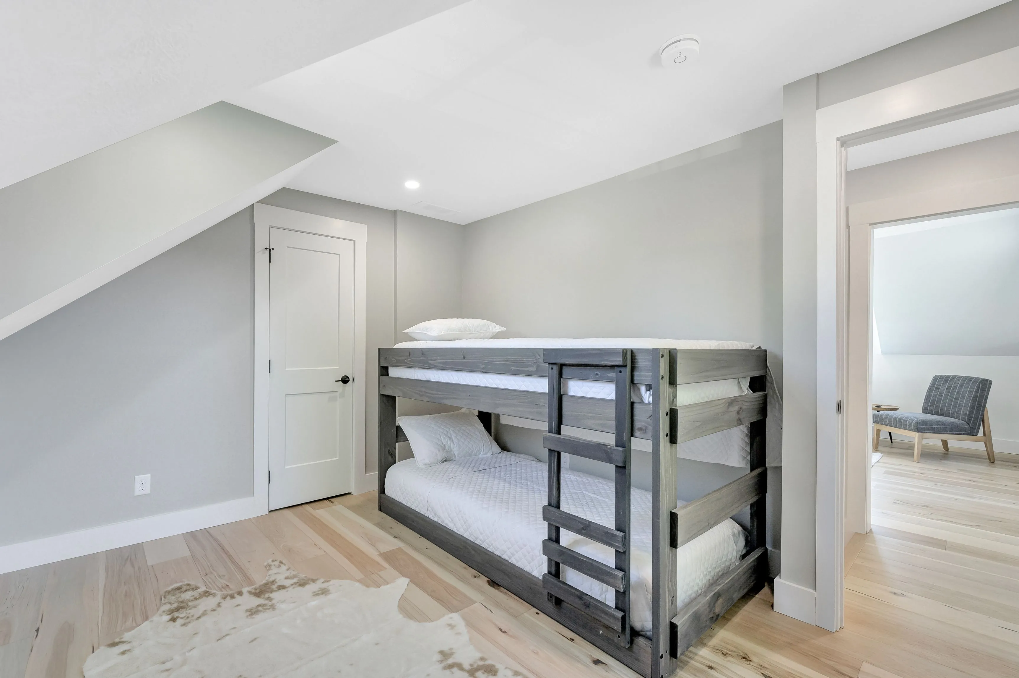 A contemporary room with a wooden bunk bed, white bedding, light hardwood floors, and a small chair by the doorway.