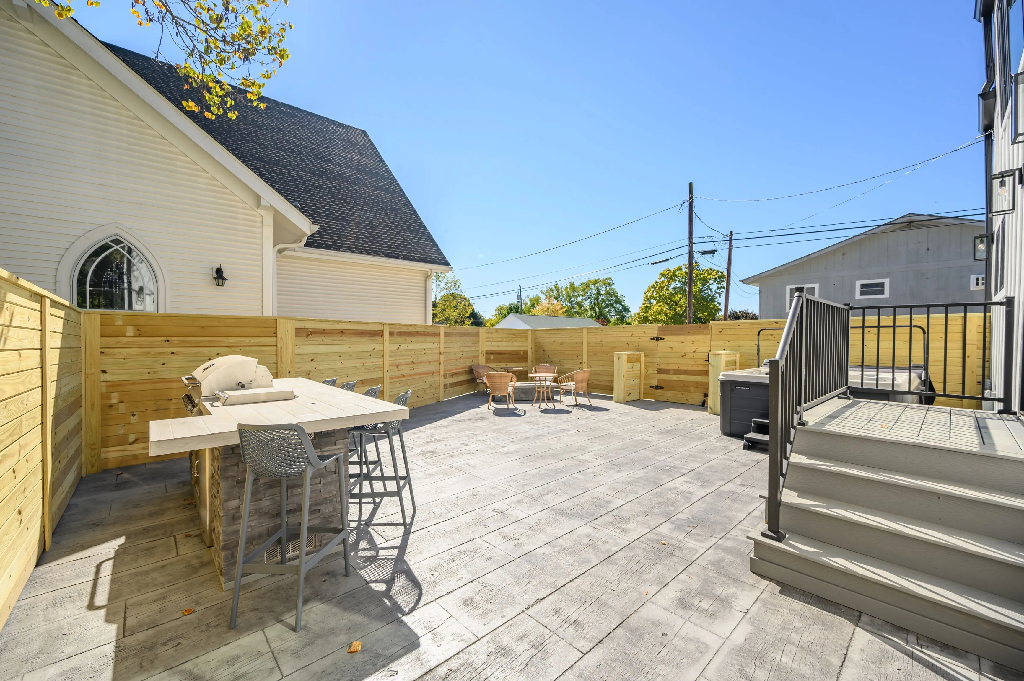 Spacious backyard patio area with wooden privacy fence, outdoor furniture, and a barbecue grill beside a residential building on a sunny day.