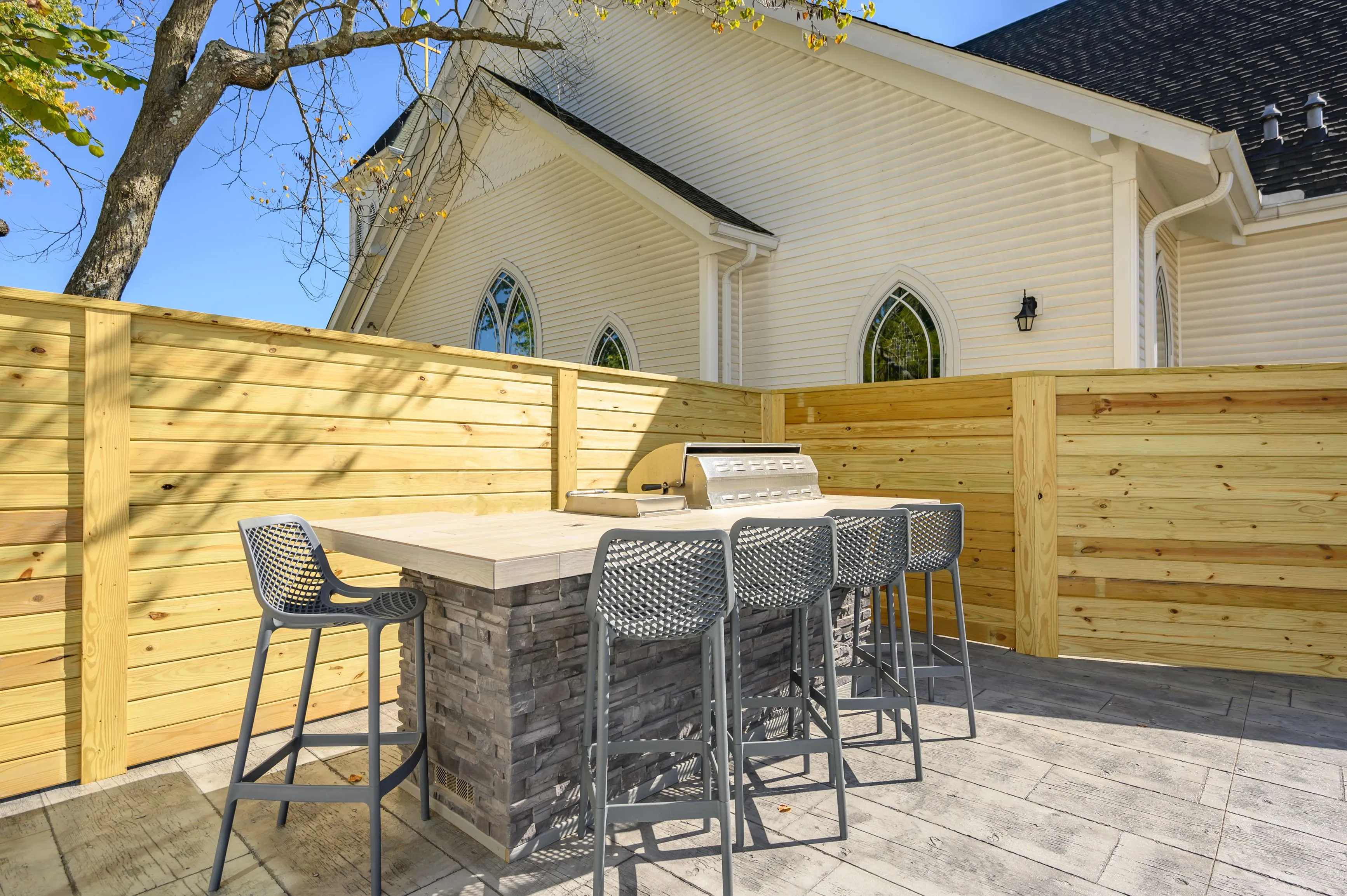 Outdoor patio with a stone bar and high stools next to a wooden privacy fence with a partial view of a church in the background.