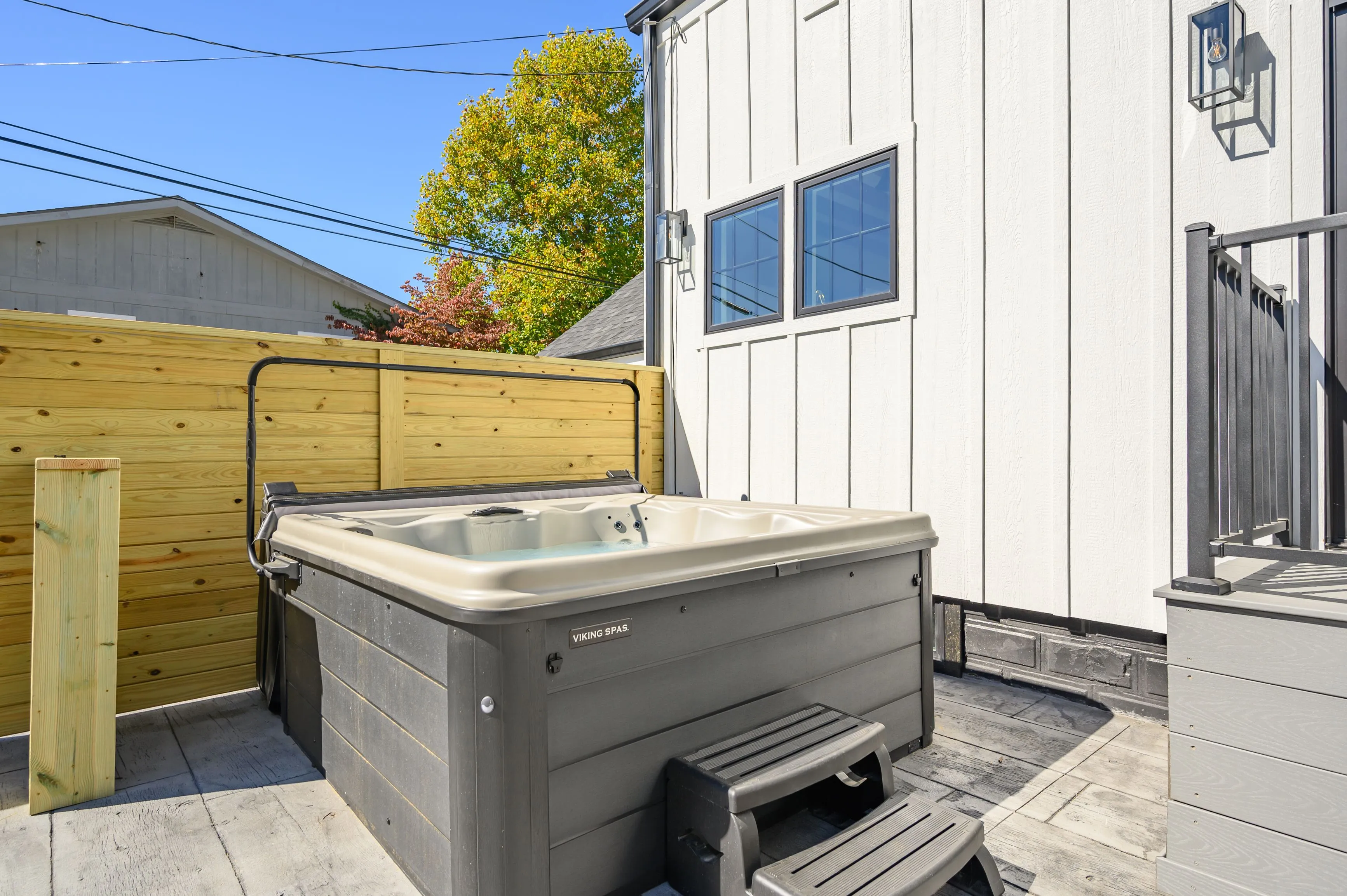 Outdoor residential hot tub with a closed cover on a wooden deck next to a wooden privacy fence, with a white exterior wall and windows in the background, on a sunny day.