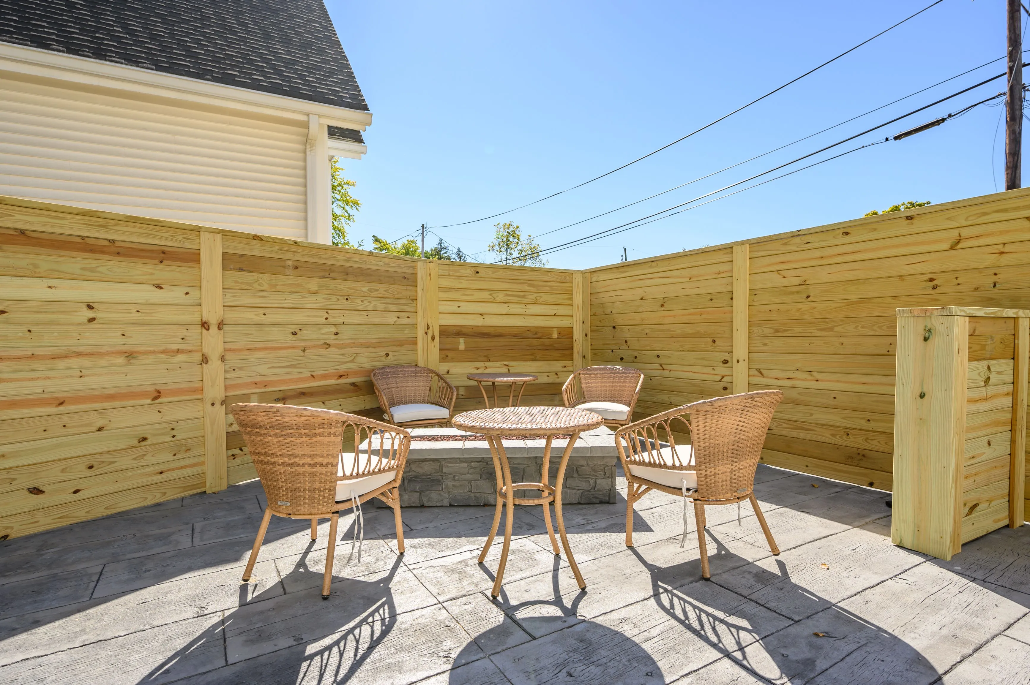 Outdoor patio area with a wooden fence, stone flooring, and a set of wicker chairs around a small round table.