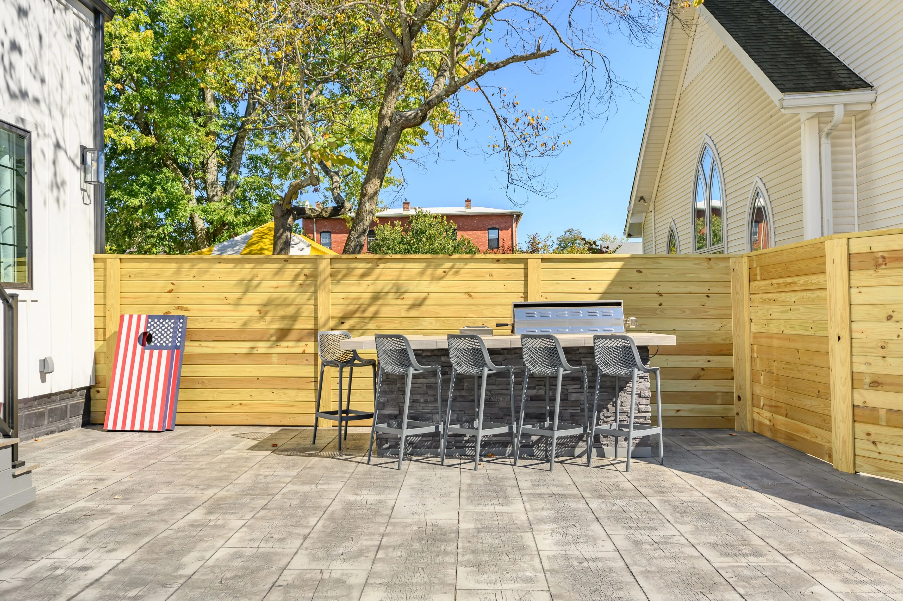 Outdoor patio area with modern furniture, a barbecue grill, and a cornhole game board, surrounded by wooden fencing with trees and a church building in the background.