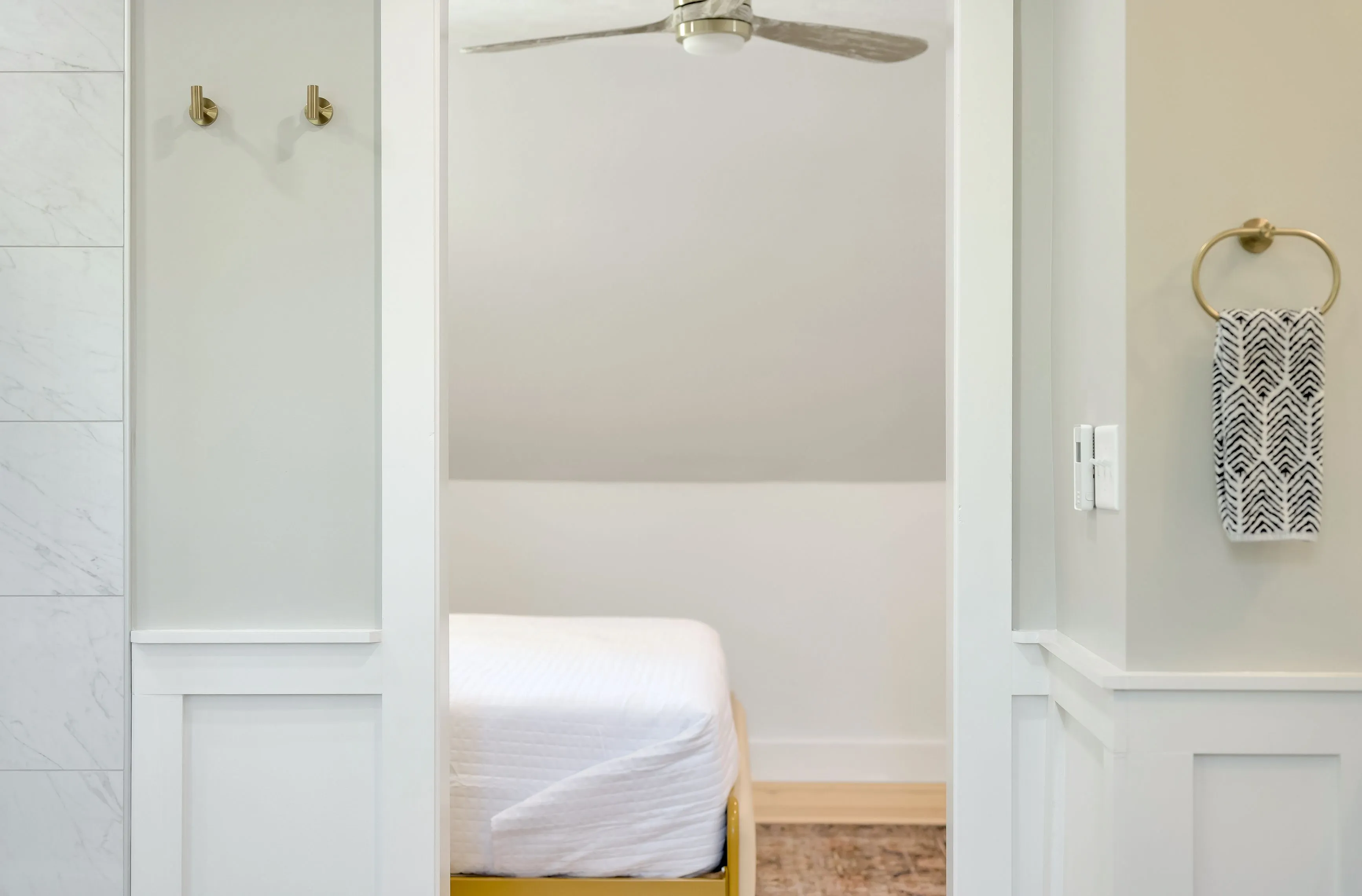 View from a bathroom looking into a bedroom with an open door, showing a ceiling fan above and a bed with white bedding in the distance.