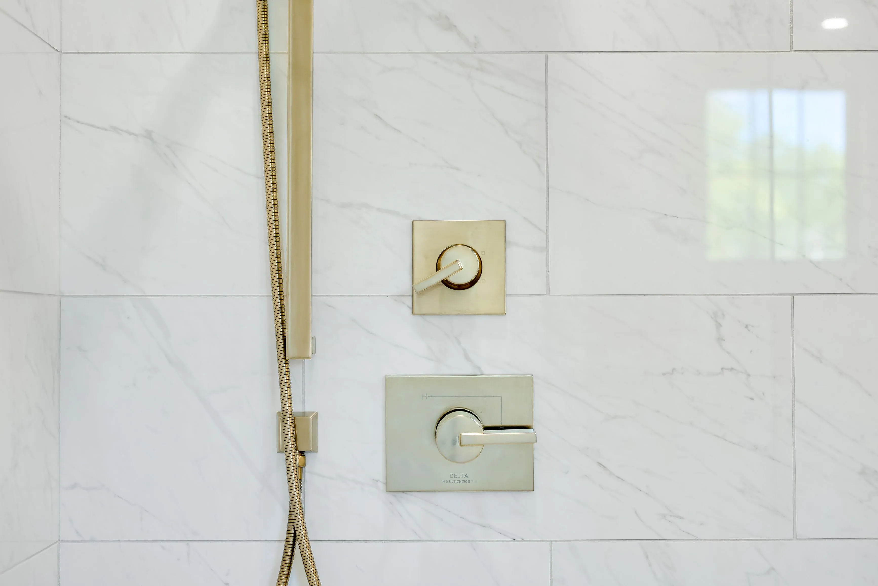 Modern bathroom wall with marble tiles featuring a gold shower handle and two temperature control valves.